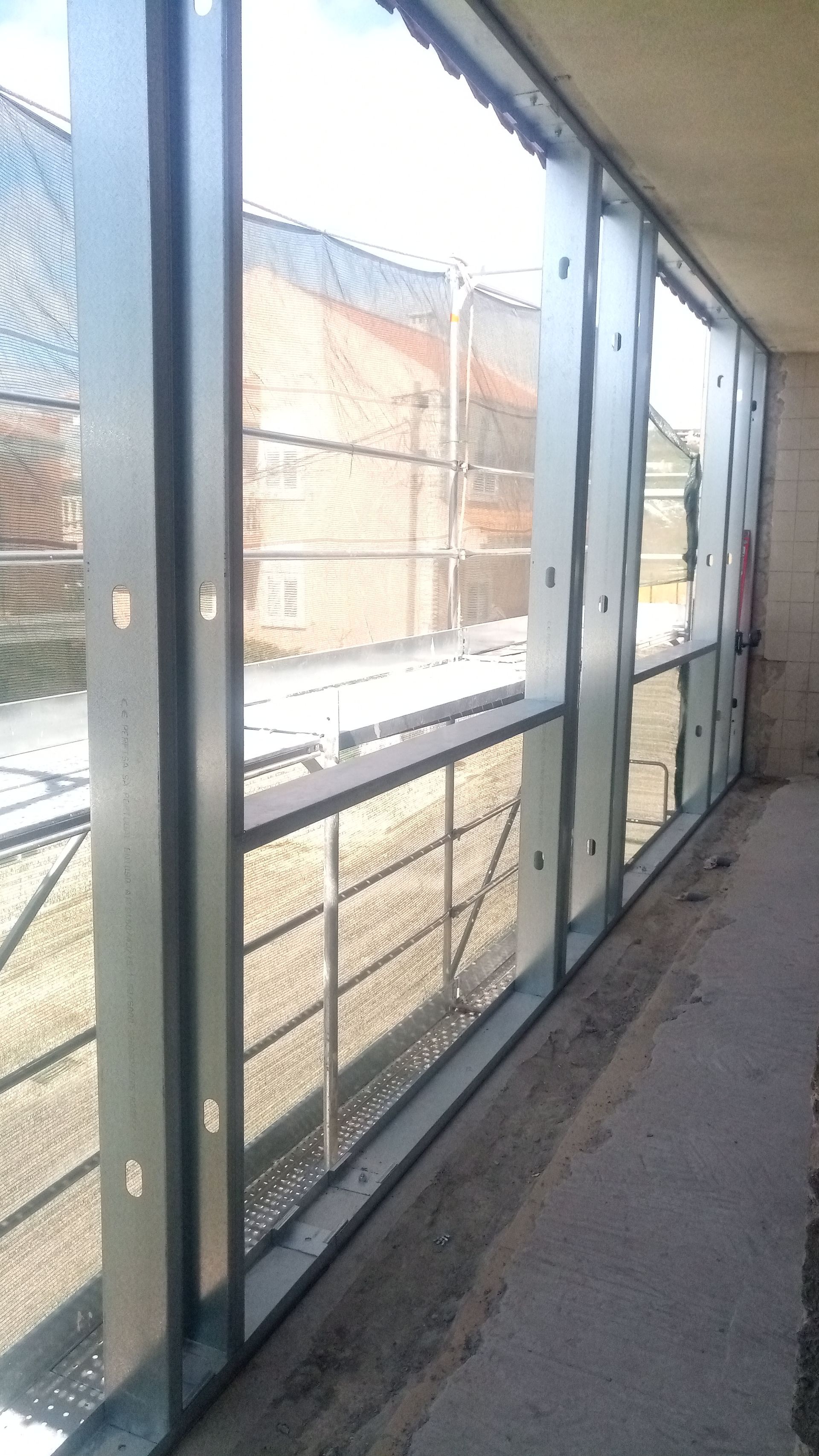 Janelas com estrutura metálica e grades de segurança com vista para uma área externa cercada por uma cerca e um prédio.