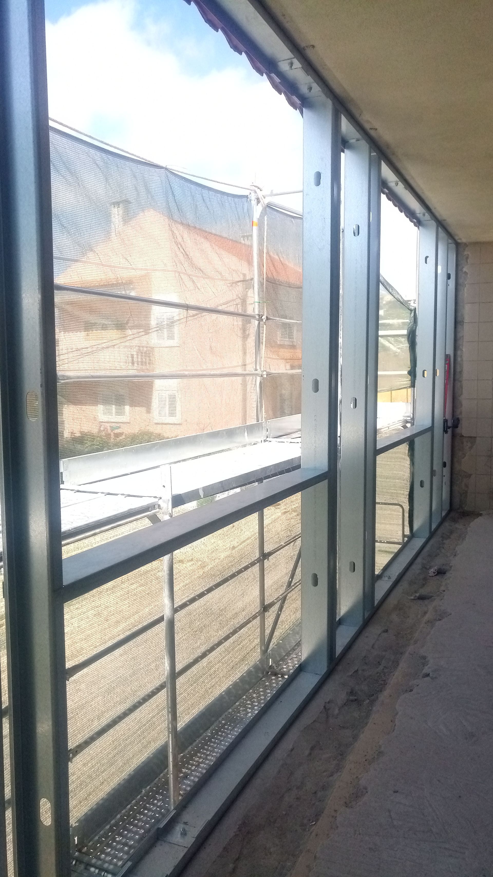 Janelas com estrutura metálica e tela de arame cobrindo uma paisagem externa que inclui um edifício e o céu.