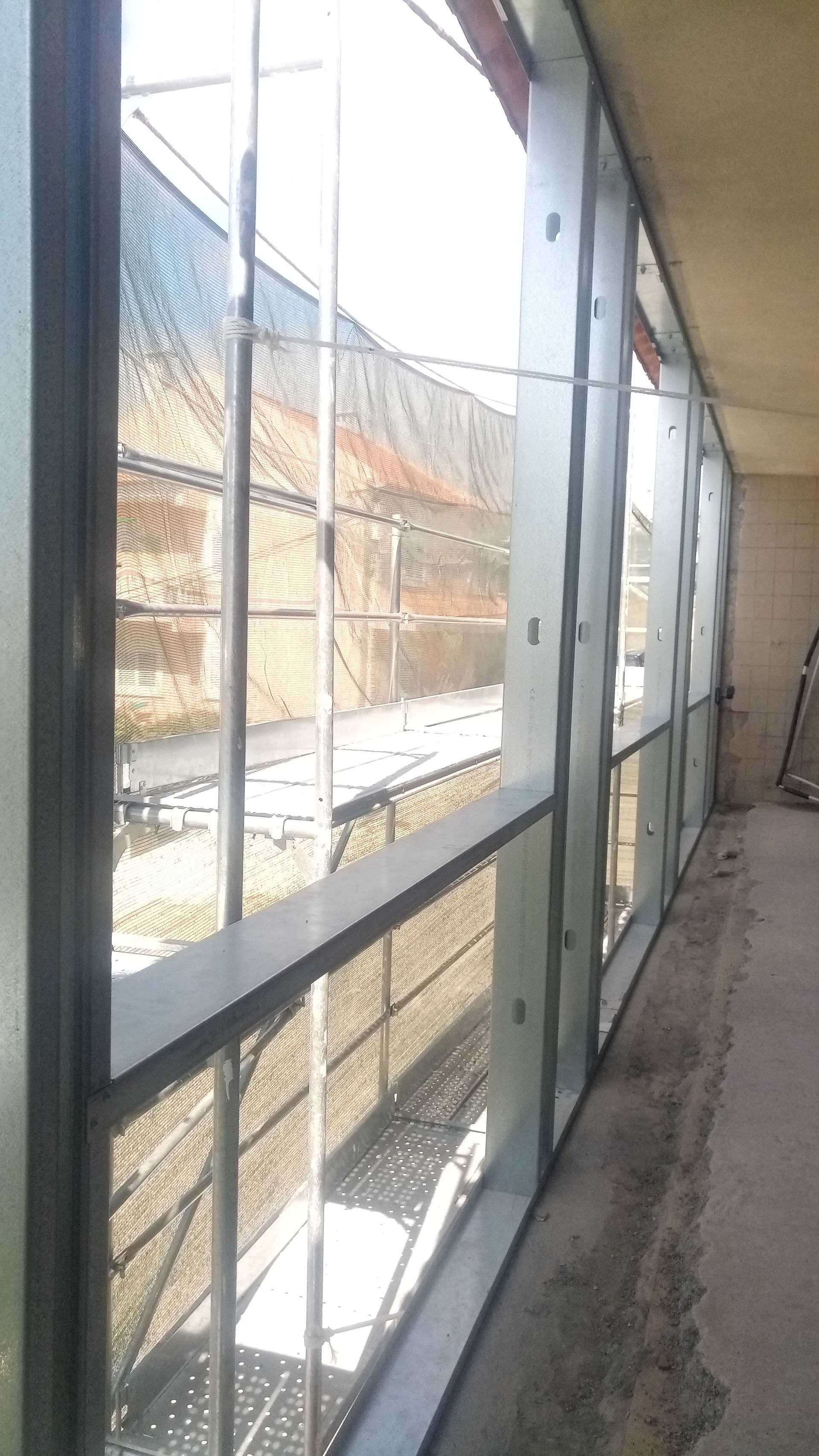 Vista exterior através de uma fileira de janelas altas. Parede de tijolos e luz solar visíveis do lado de fora.