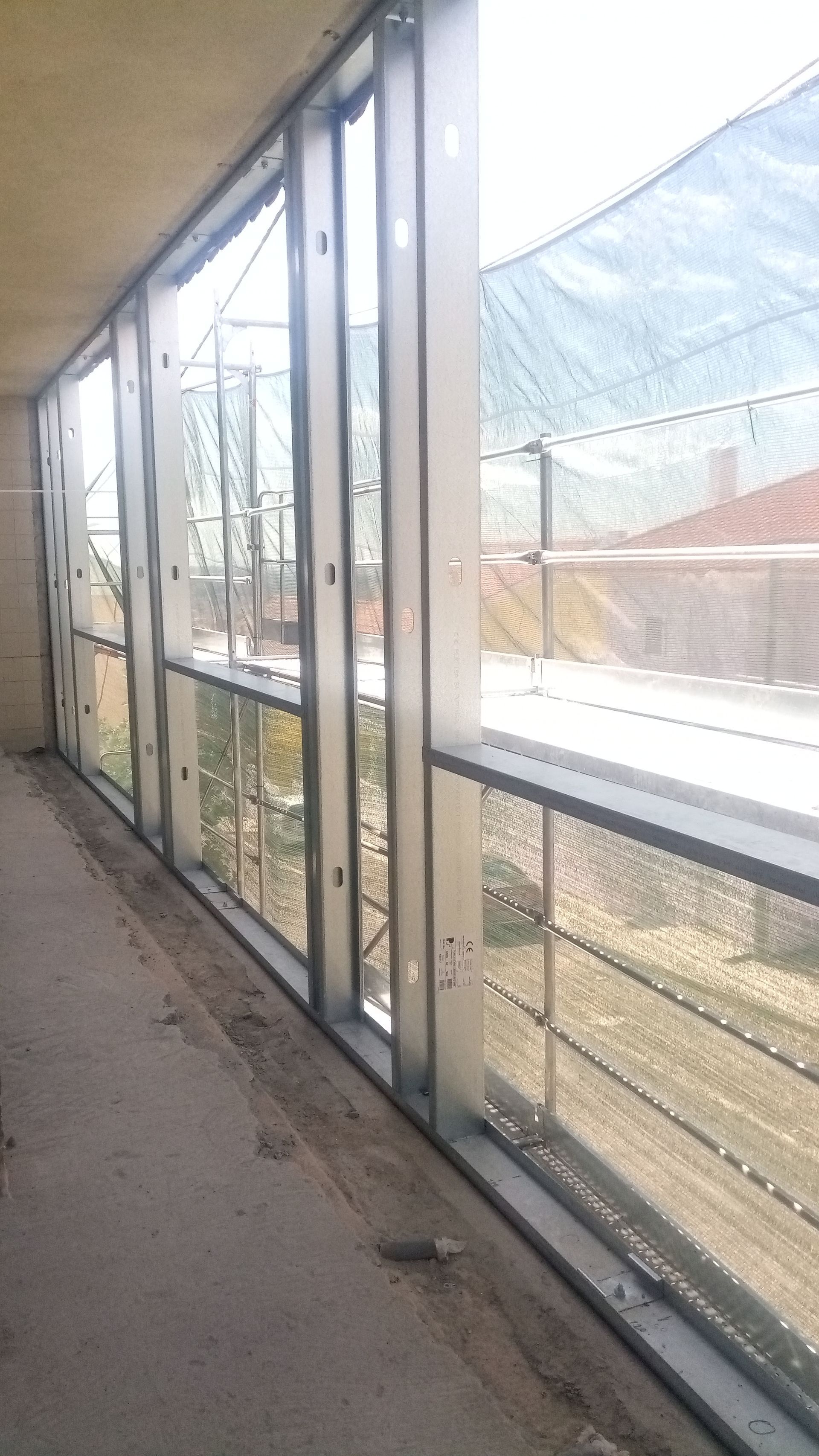 Janelas com moldura de metal ao longo de uma parede. Canteiro de obras visível do lado de fora, com andaimes e lona.