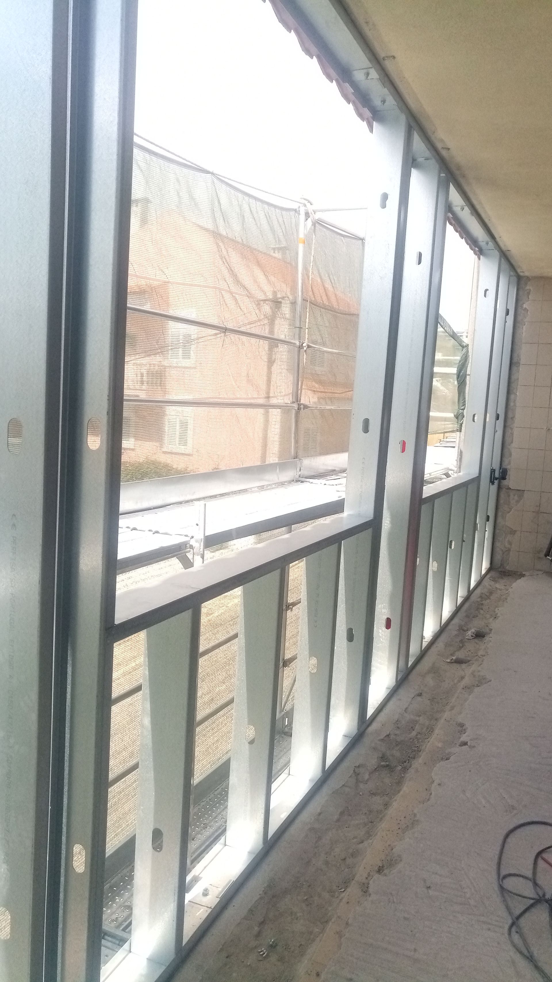 Vista interior de um edifício em construção, com janelas de caixilharia metálica e vista para outros edifícios e uma cerca no exterior.