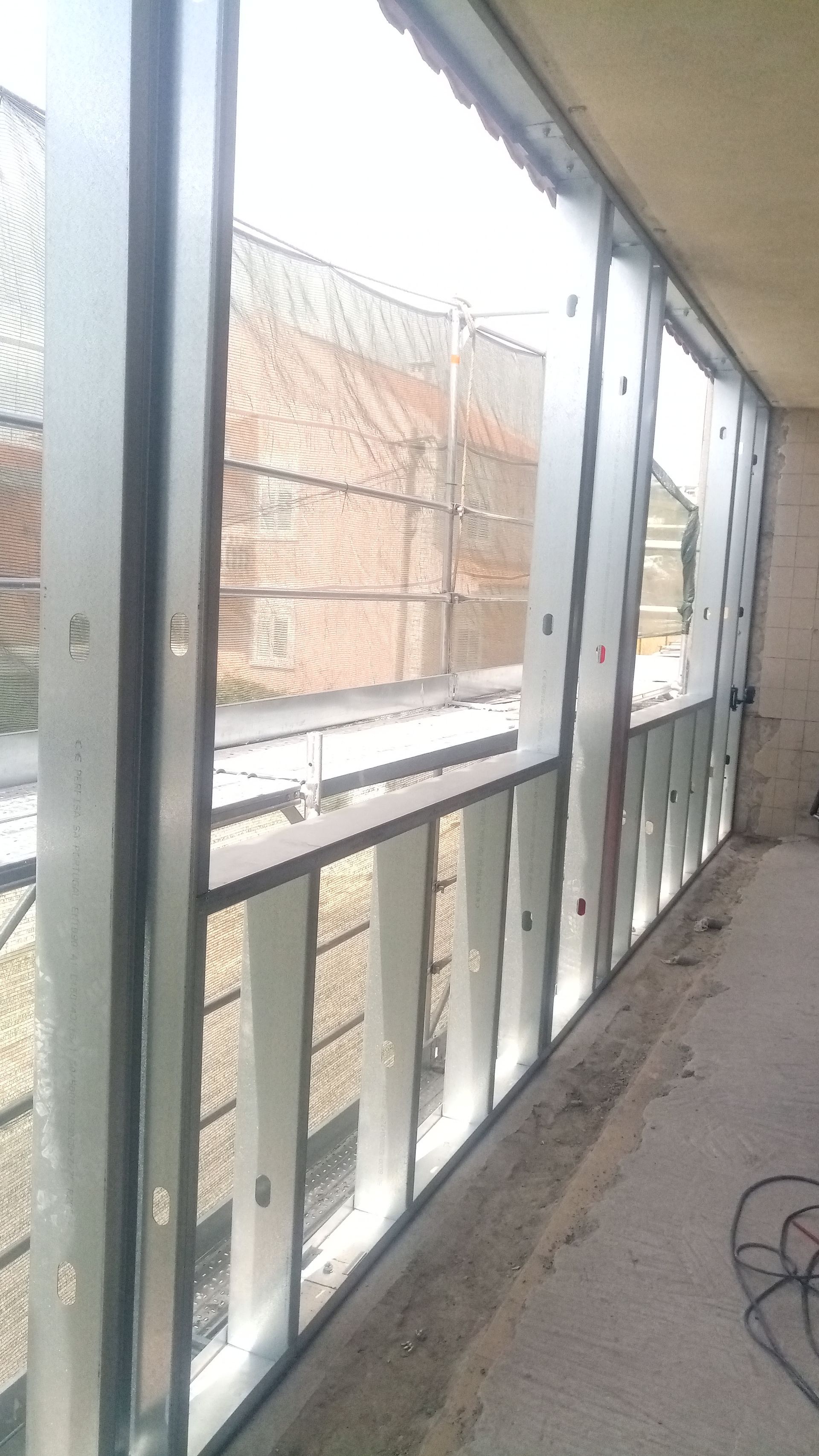 Caixilhos de metal instalados, mostrando o interior inacabado de um edifício com vista para uma estrutura de tijolos do lado de fora.