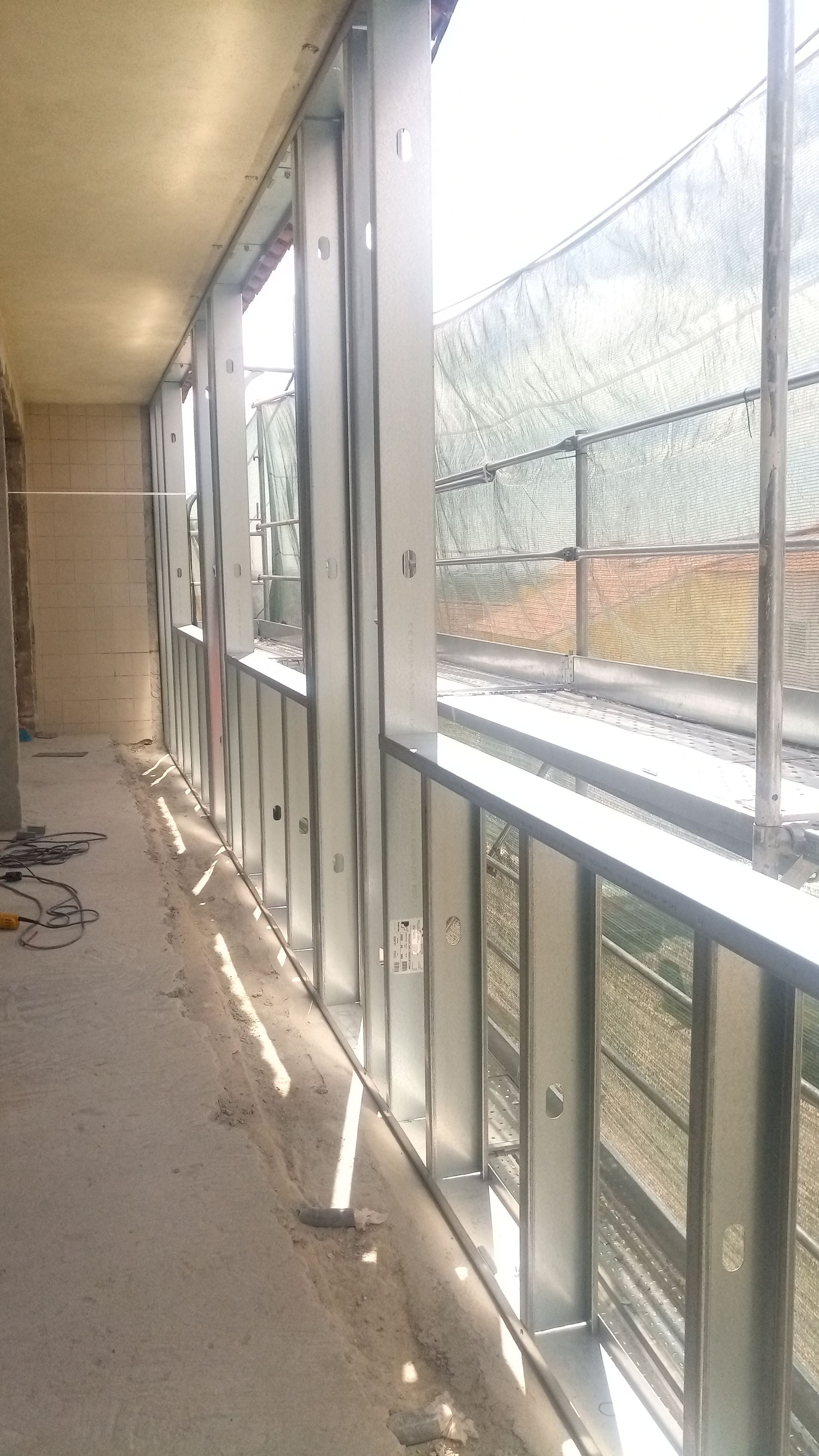 Estrutura metálica de uma janela e grade ao longo de um corredor de concreto, com uma cerca visível do lado de fora.