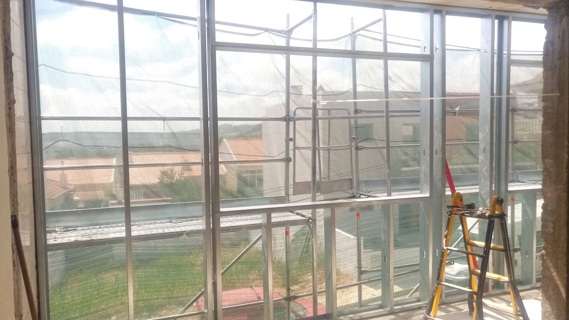 Janela com estrutura metálica e tela mosquiteira, parcialmente construída, com vista para edifícios e árvores.