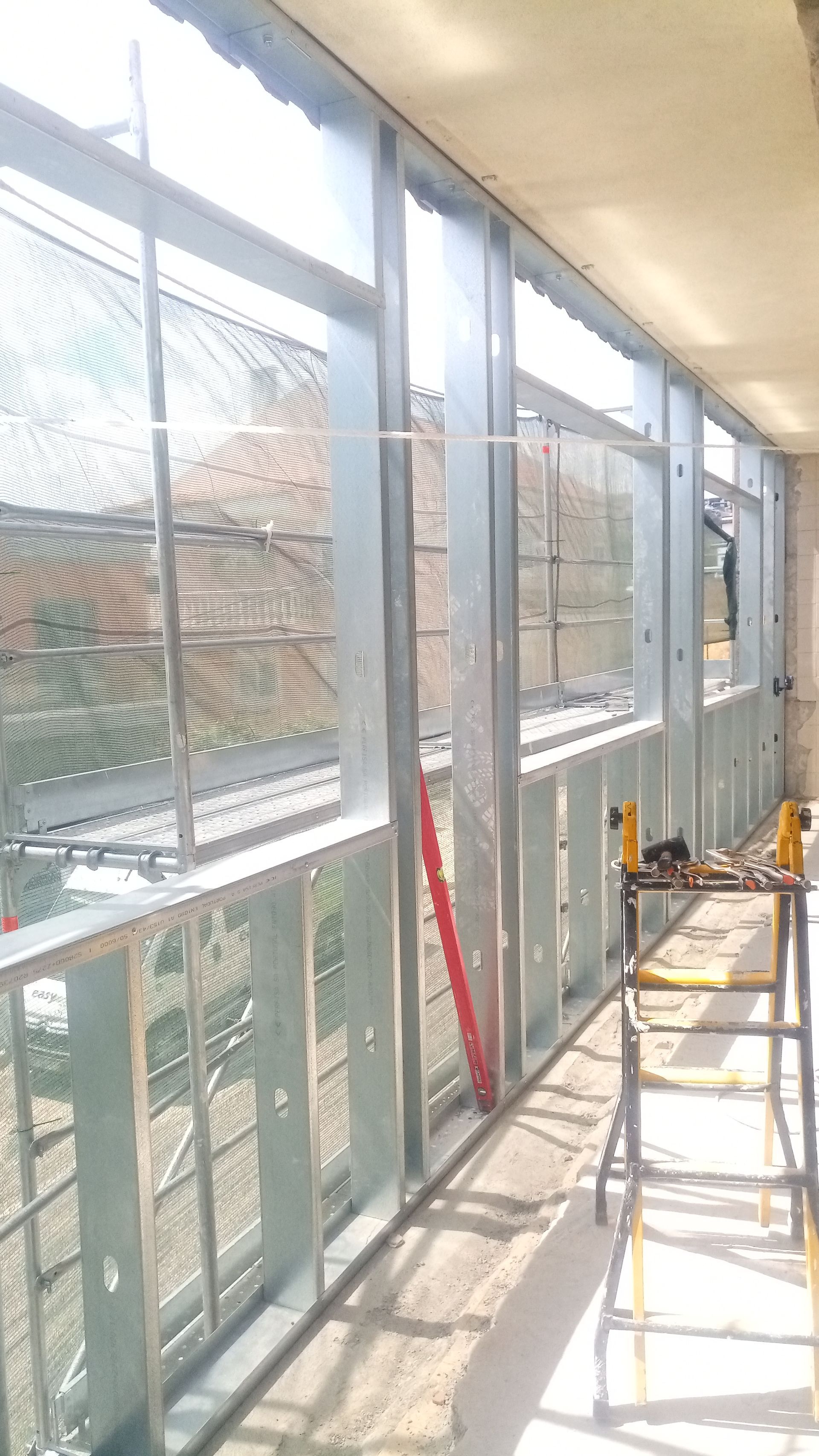 Estrutura metálica de uma parede e janela, em um canteiro de obras. Uma escada está em primeiro plano.