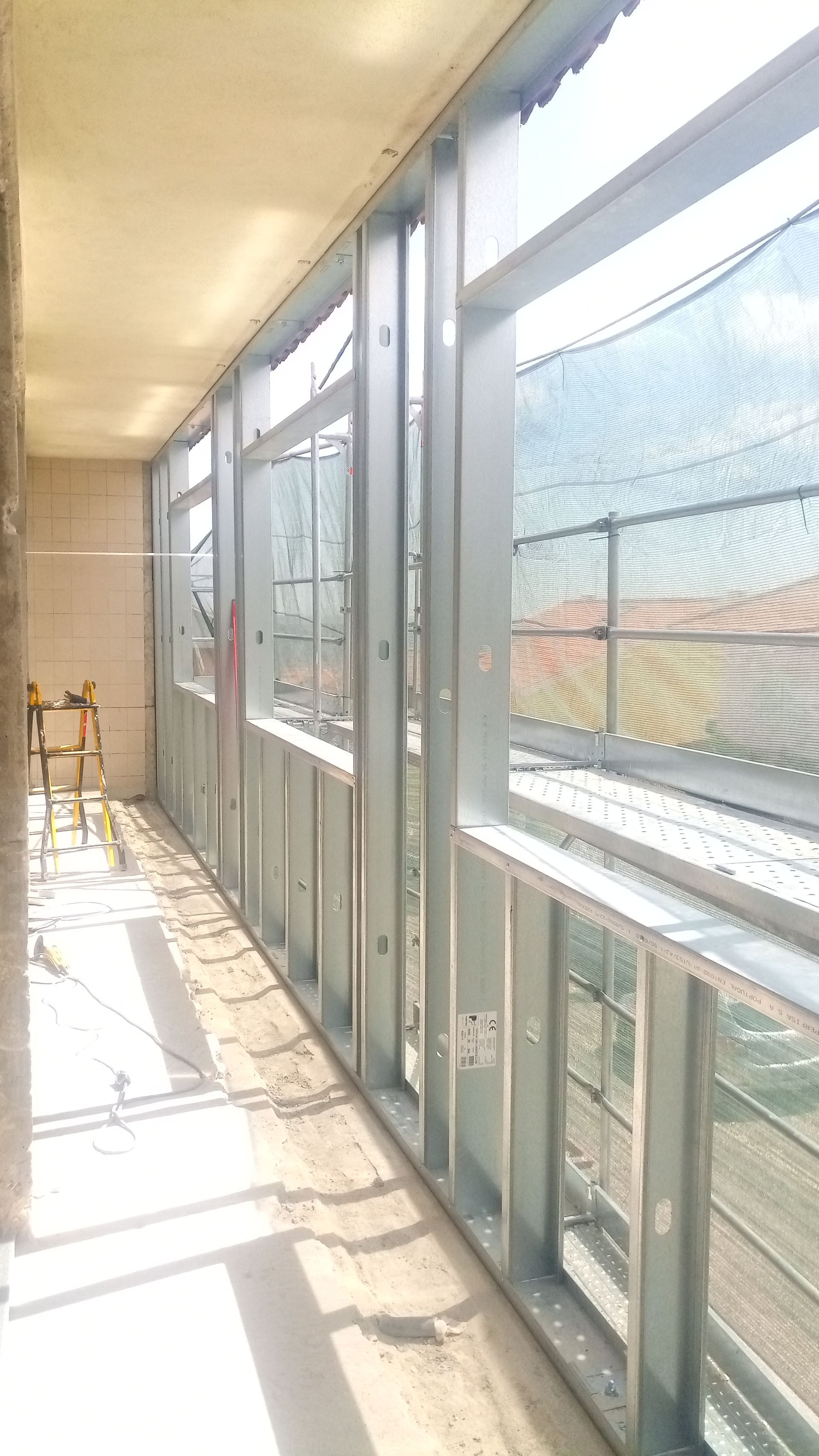 Vista interna de um canteiro de obras. Estrutura de aço ao longo de uma janela, paredes inacabadas e vista para o exterior.