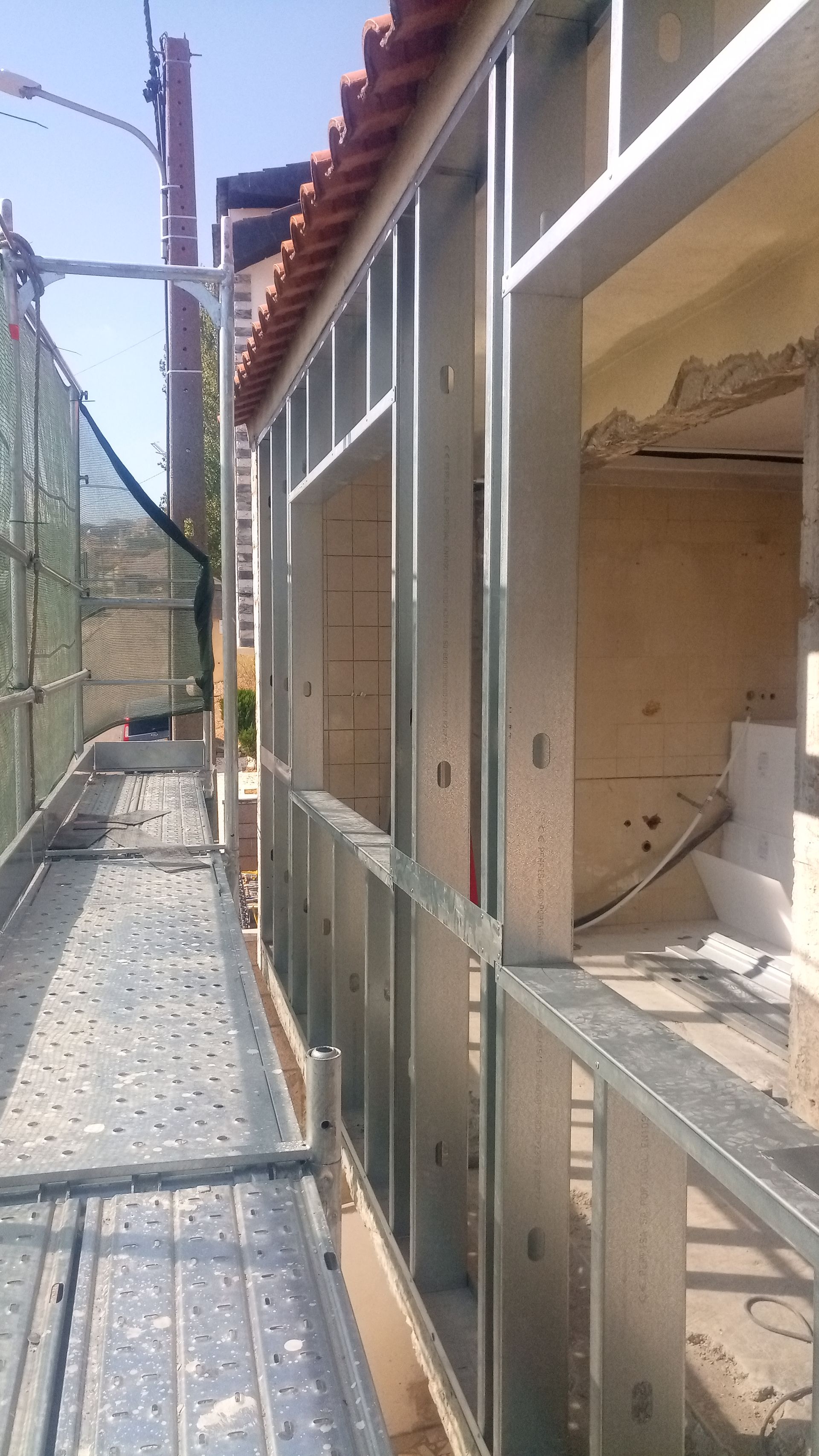 Estrutura metálica da parede externa de um edifício. Andaimes visíveis. Telhado de telhas vermelhas.