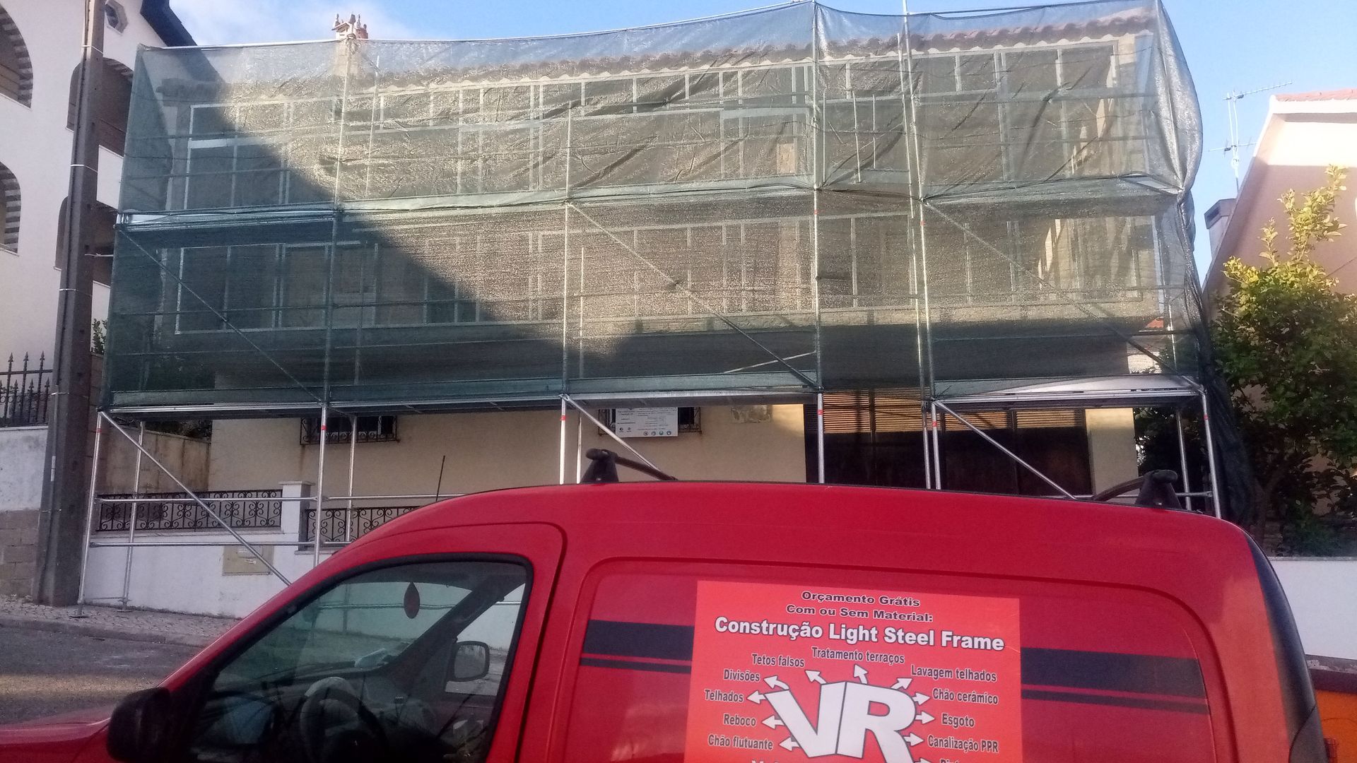 Prédio em construção, com andaimes e coberto por uma rede verde, com uma van vermelha em primeiro plano.