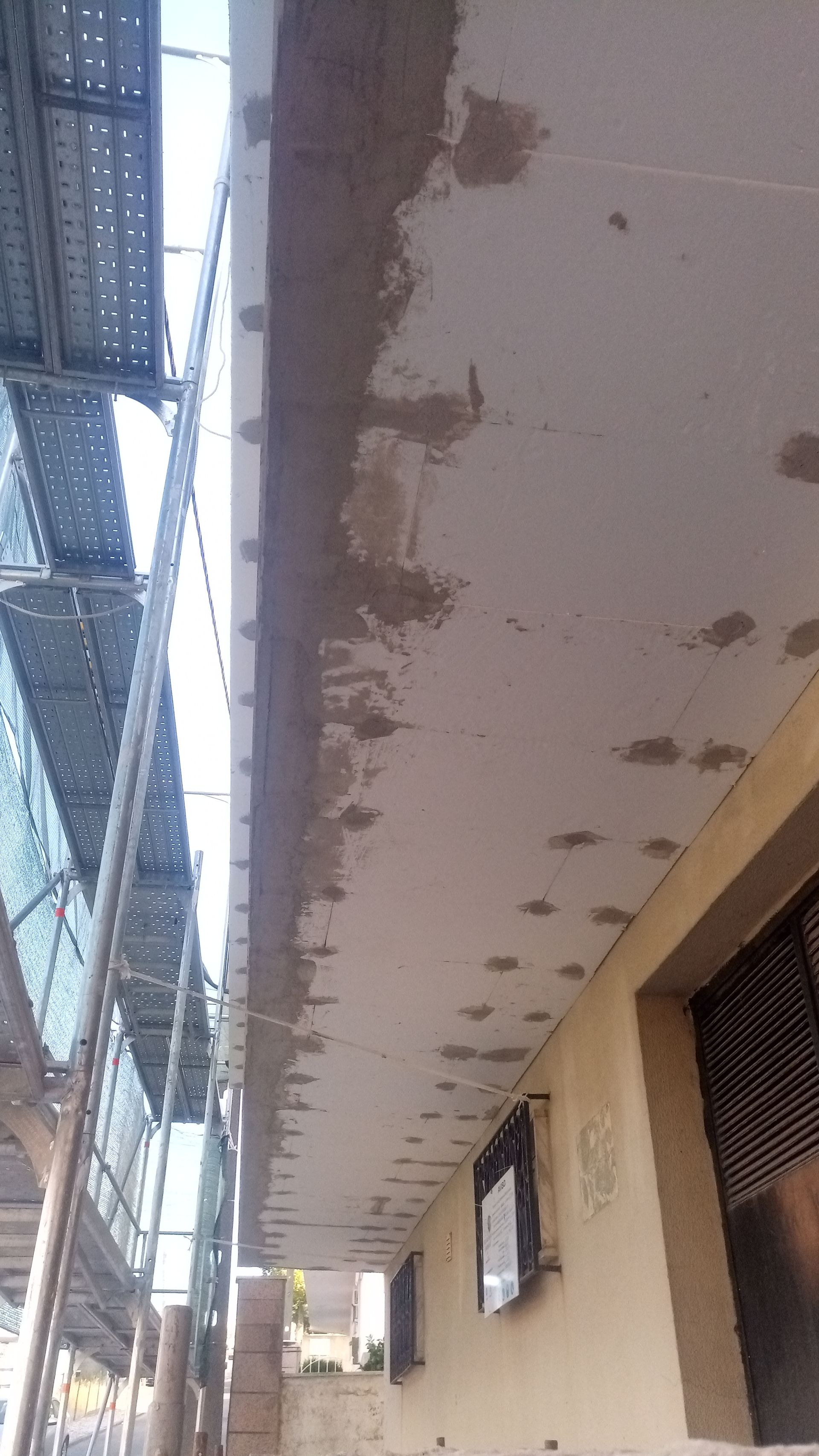 Andaimes montados ao longo da parte inferior de um edifício com gesso fresco aplicado em seções; luz solar.