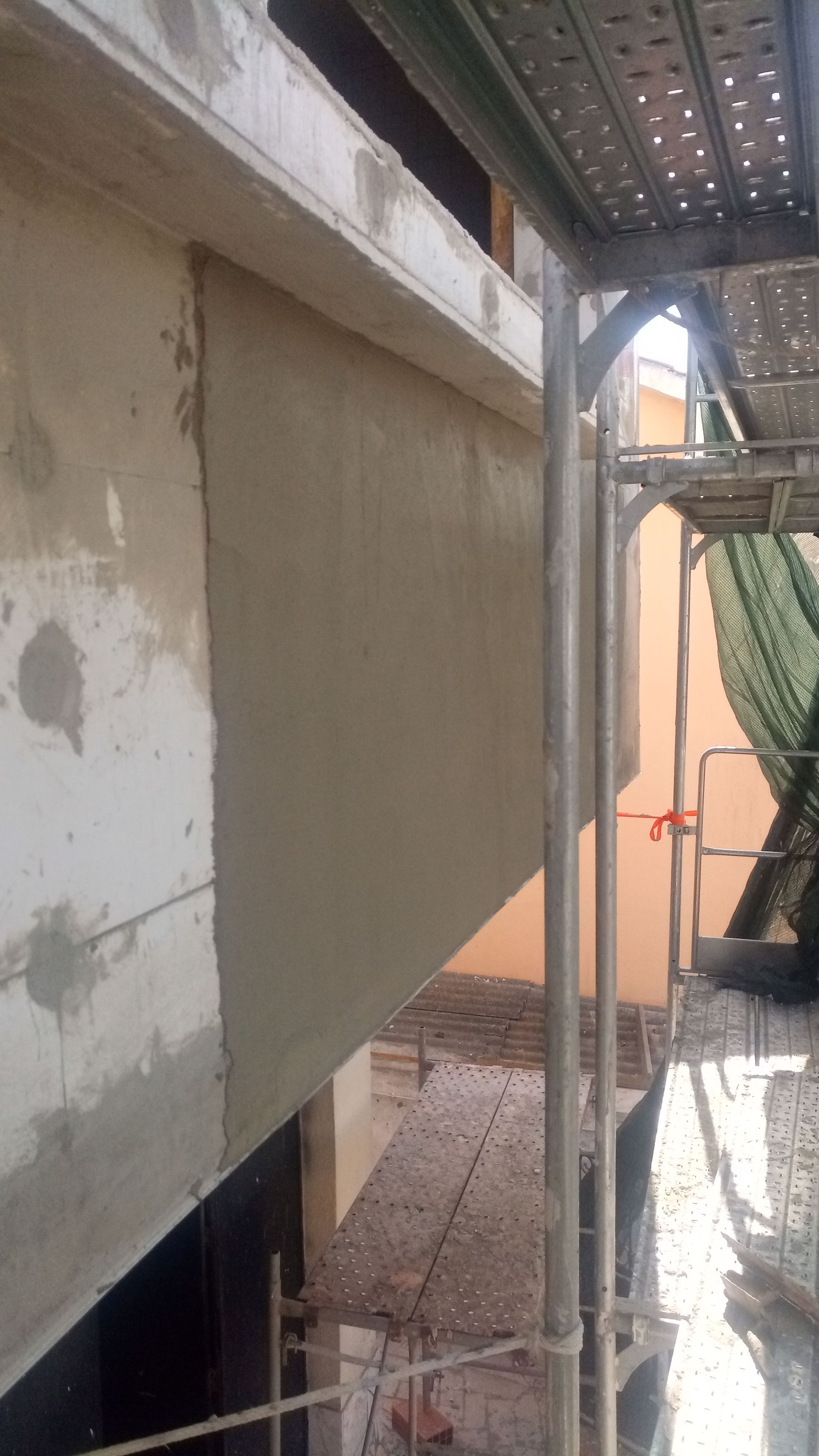 Parede externa do edifício durante a construção, com andaimes à direita, rebocada com cimento cinza.