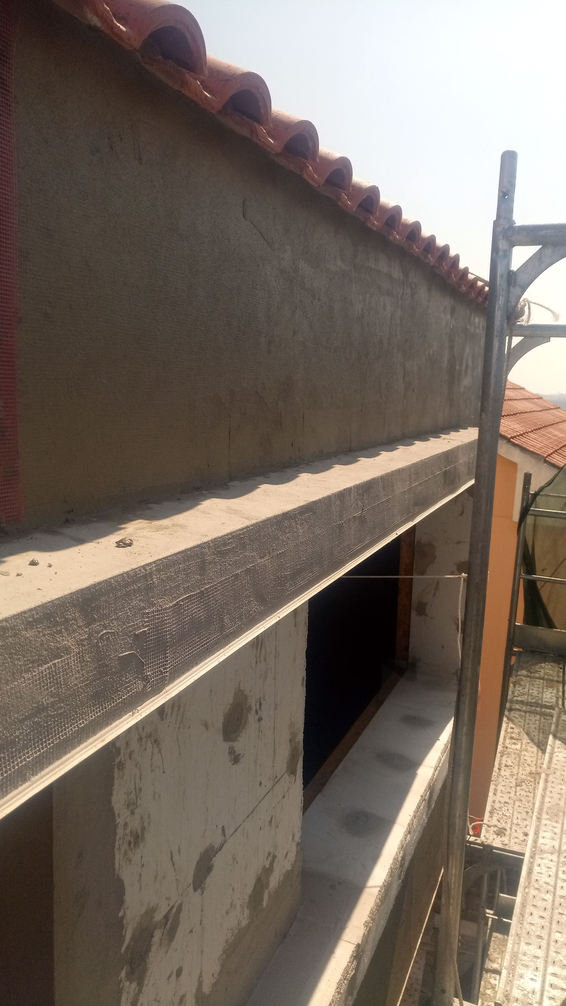 Prédio em construção, paredes de estuque marrom, telhado de telha vermelha, andaimes ao lado.