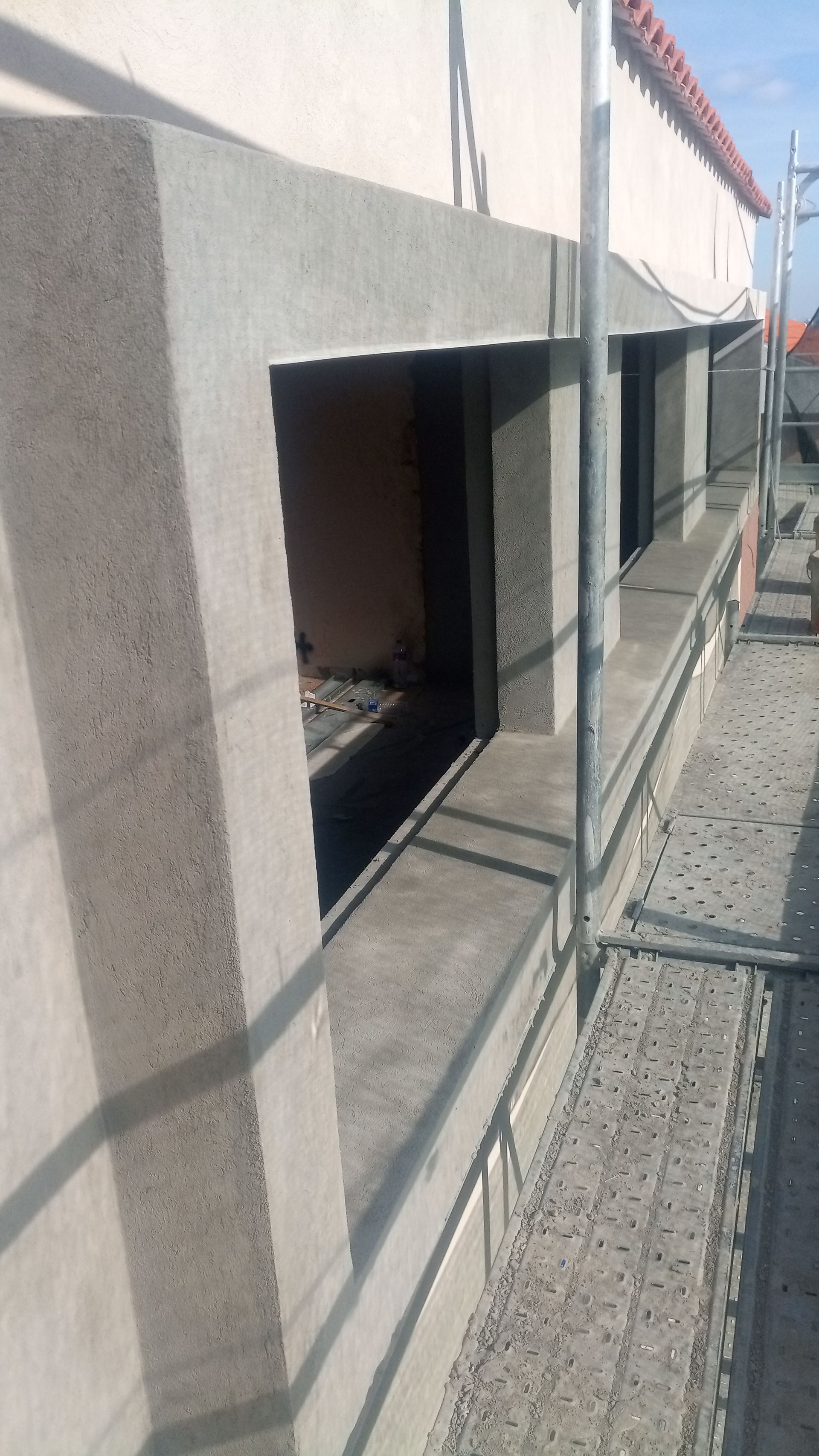 Parede externa com aberturas para janelas, caixilhos de cimento cinza, andaimes de construção e telhado avermelhado.