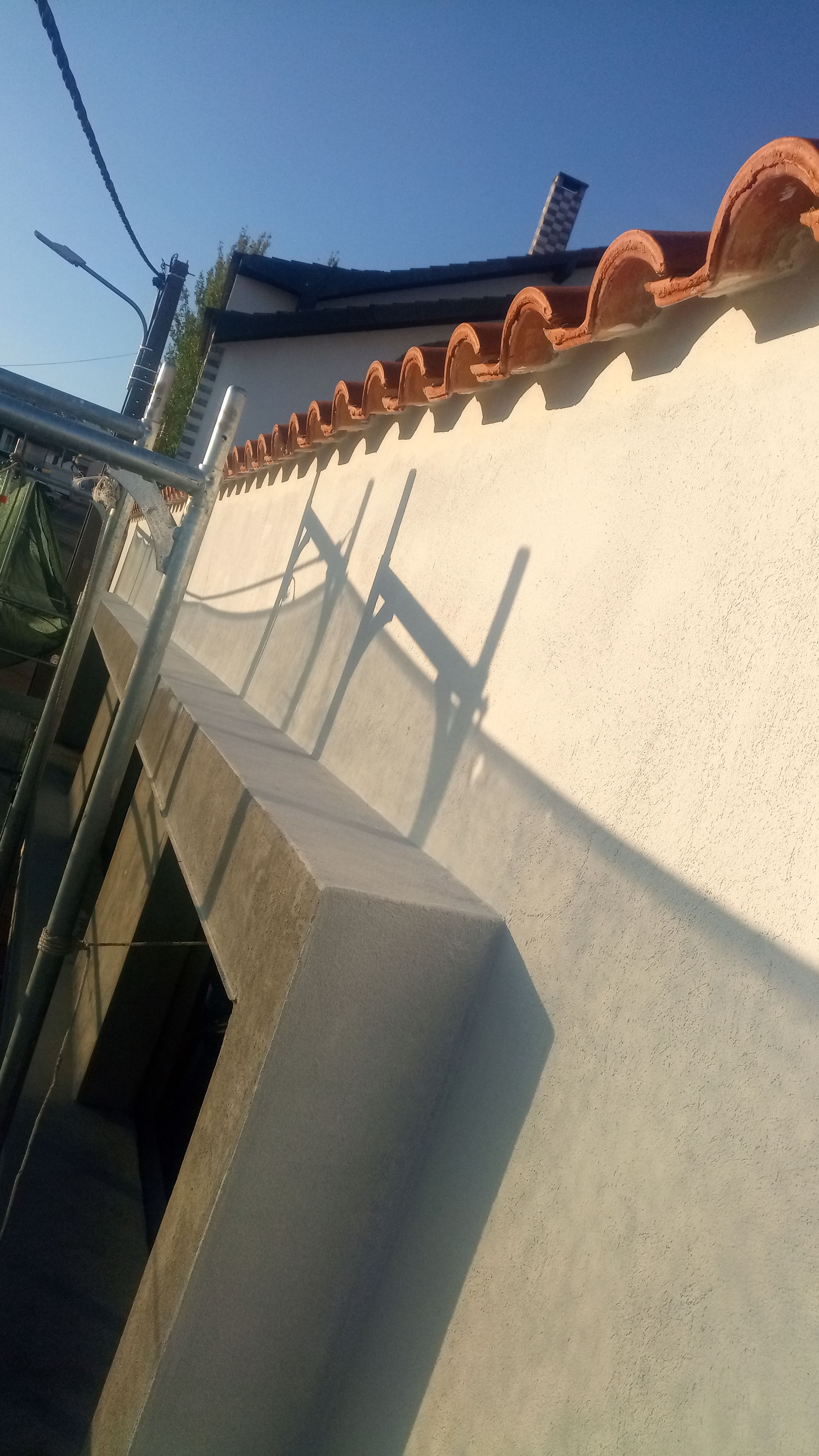 Exterior de um edifício com telhas de terracota, paredes de estuque de cor clara e andaimes de construção projetando uma sombra.