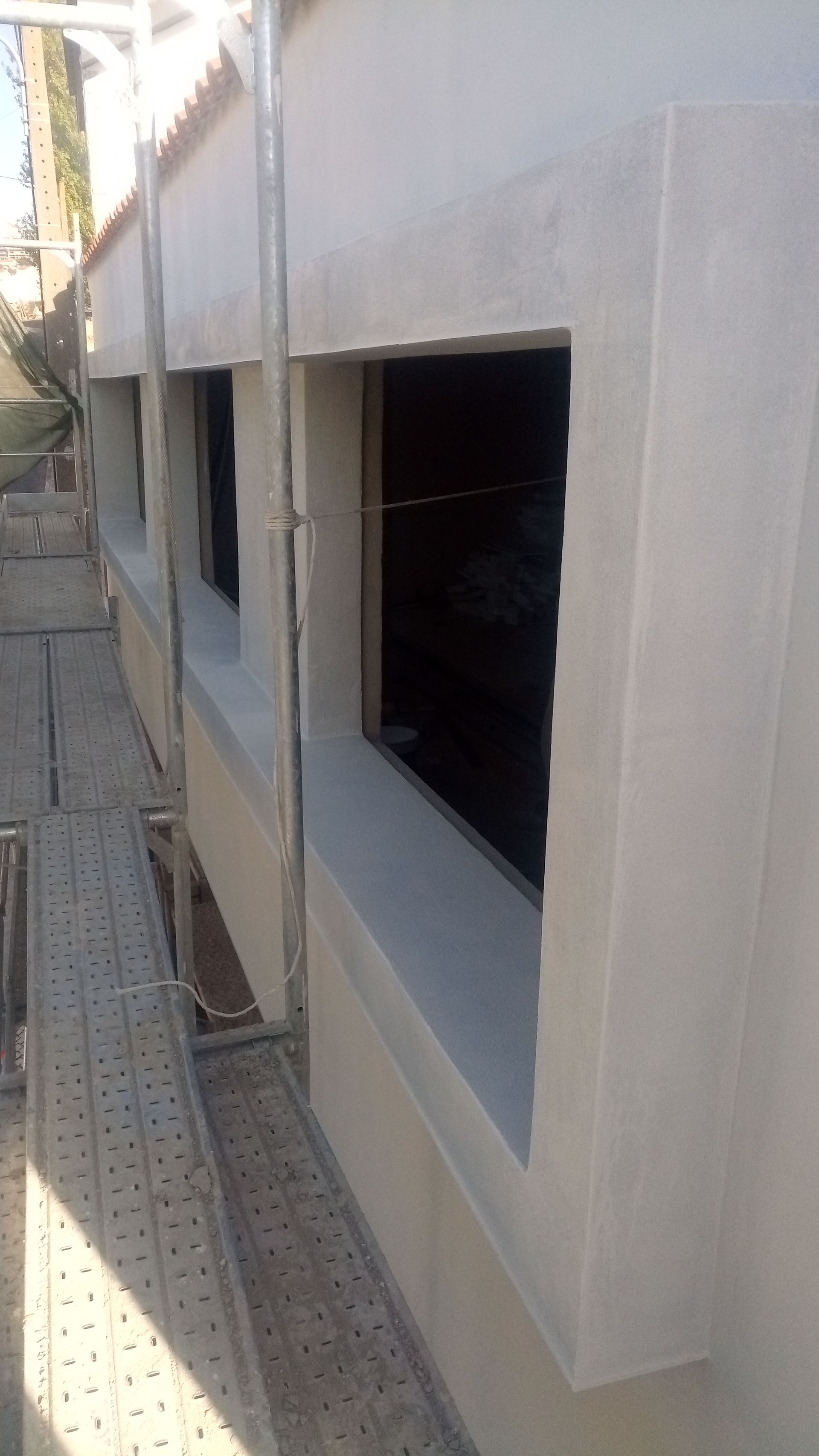 Parede externa com três caixilhos de janelas vazios; um andaime metálico está montado ao lado.