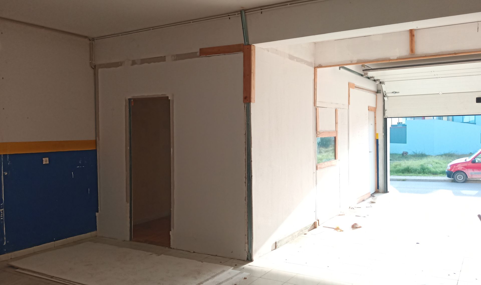 Interior de um prédio comercial. Apresenta uma pequena sala com uma porta aberta, uma parede pintada de azul e amarelo e uma grande porta de garagem aberta.
