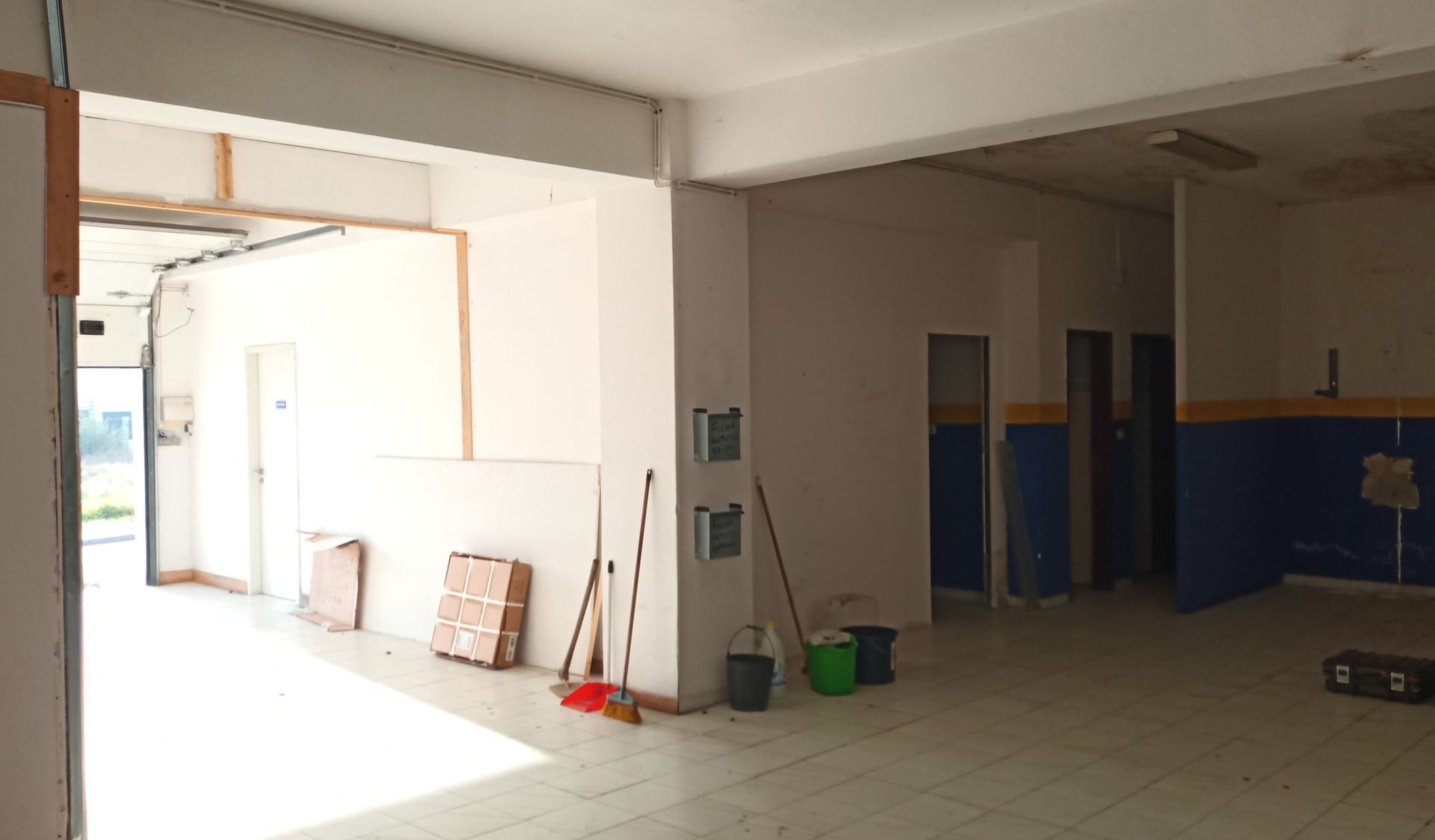 Interior vazio de um edifício com uma grande abertura, paredes brancas e piso de azulejos; materiais de construção estão presentes.