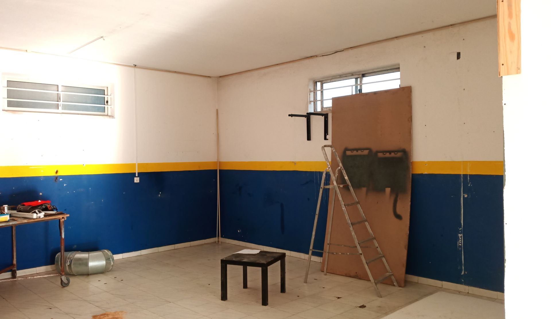 Sala vazia com paredes azuis e amarelas, uma pequena mesa, uma escada e uma grande tábua de madeira.