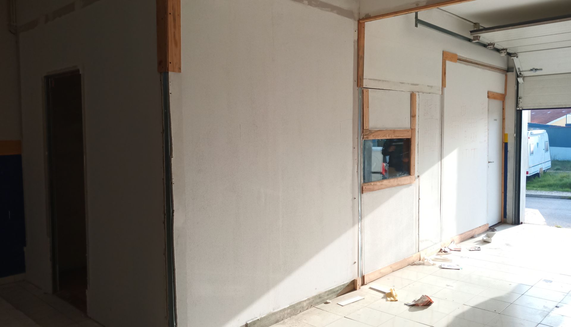 Vista interior de um cômodo em construção com paredes brancas, molduras de madeira e uma porta de garagem aberta.