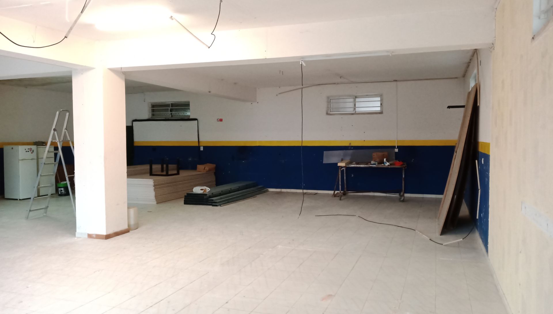 Sala vazia em tons de branco e azul com uma tela de projeção, uma escada e uma mesa.