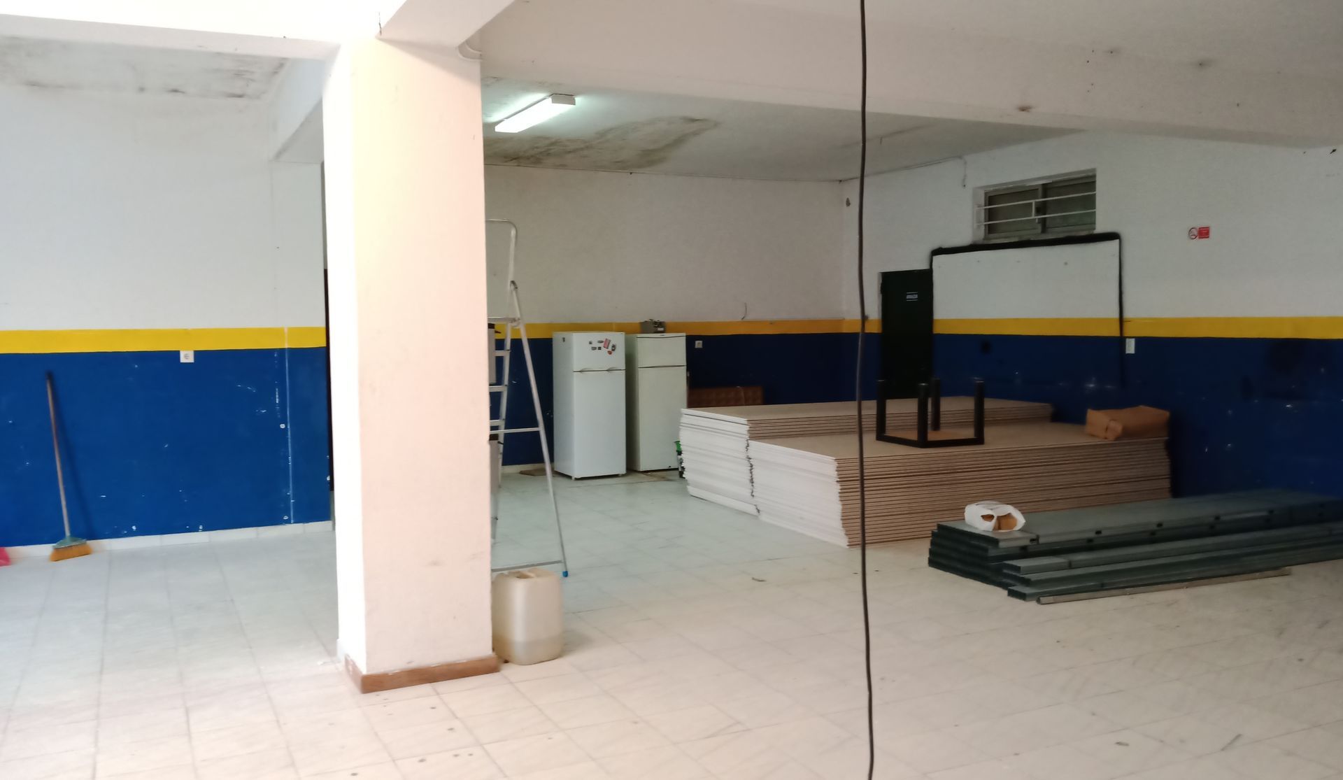 Interior de um armazém vazio com paredes pintadas de azul e amarelo, um pilar central e suprimentos.