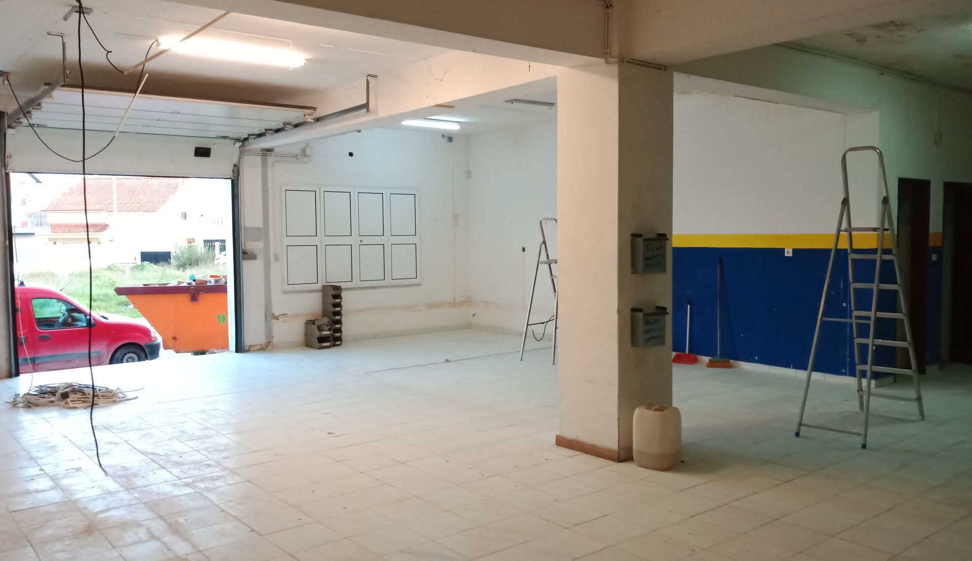 Espaço interior vazio com a porta da garagem aberta, carro vermelho visível. Paredes brancas, piso de azulejo, escada e uma seção de parede azul e amarela.