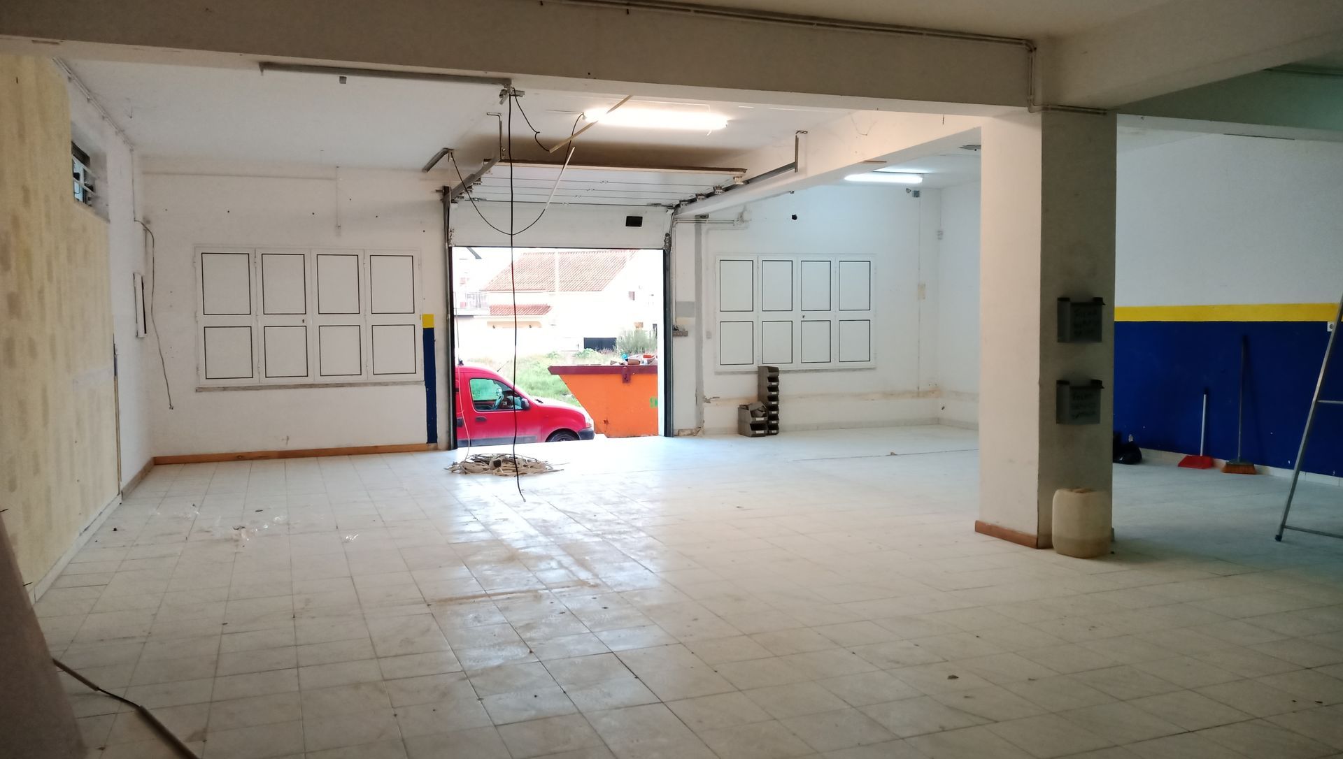 Interior de um armazém vazio com uma porta parcialmente aberta, mostrando um carro do lado de fora.