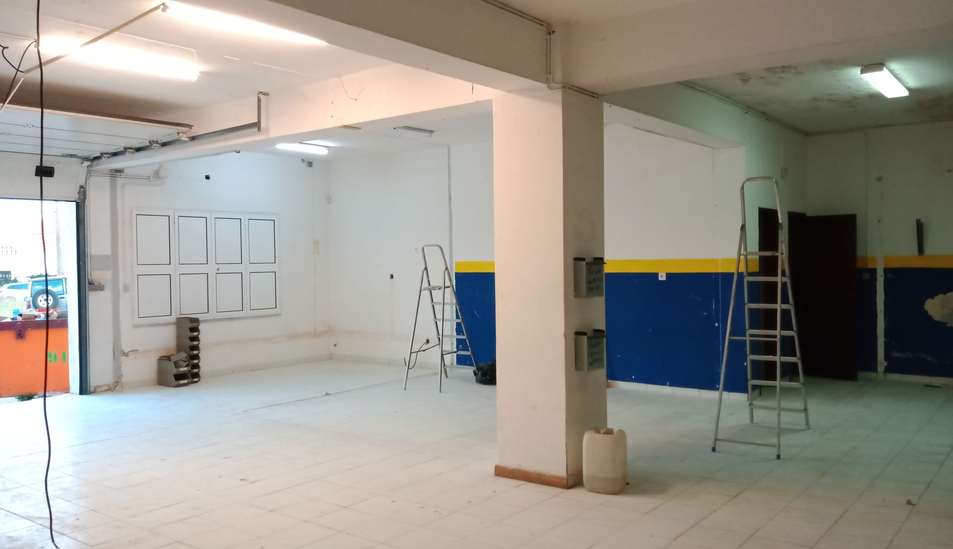 Interior vazio de uma sala com paredes brancas, detalhes em azul e amarelo, escadas e um pilar.