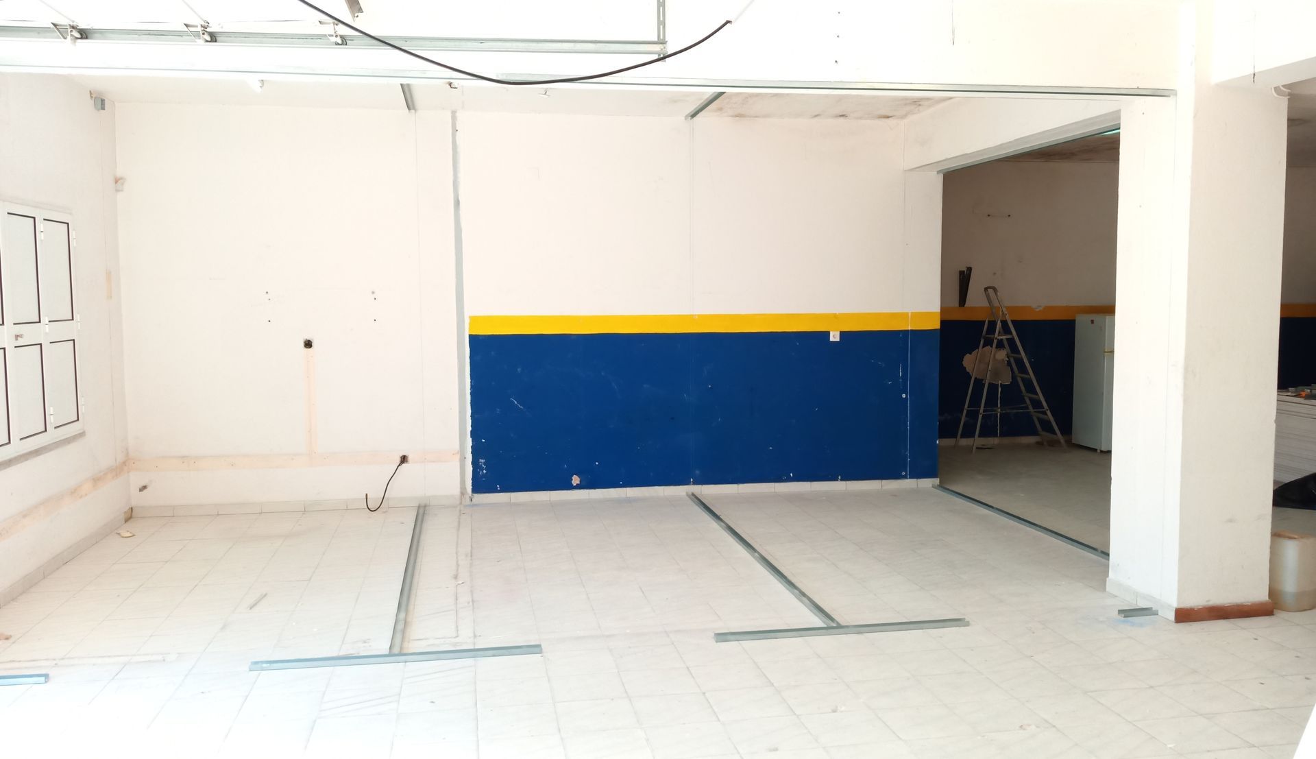Sala vazia com parede pintada de azul e amarelo, paredes brancas e piso de azulejo, possivelmente em construção.