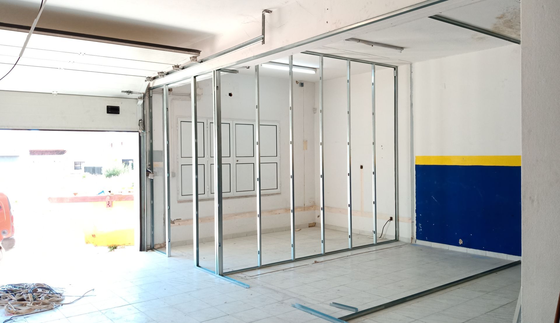 Interior com estrutura metálica parcialmente construída para uma nova parede; a porta da garagem está aberta para o exterior.