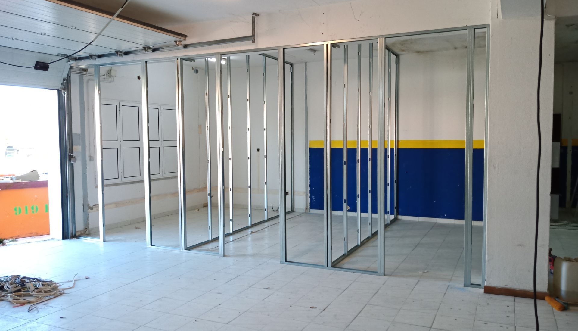 Perfis metálicos estruturam as paredes internas de um cômodo em construção. As paredes estão parcialmente pintadas de azul e amarelo.