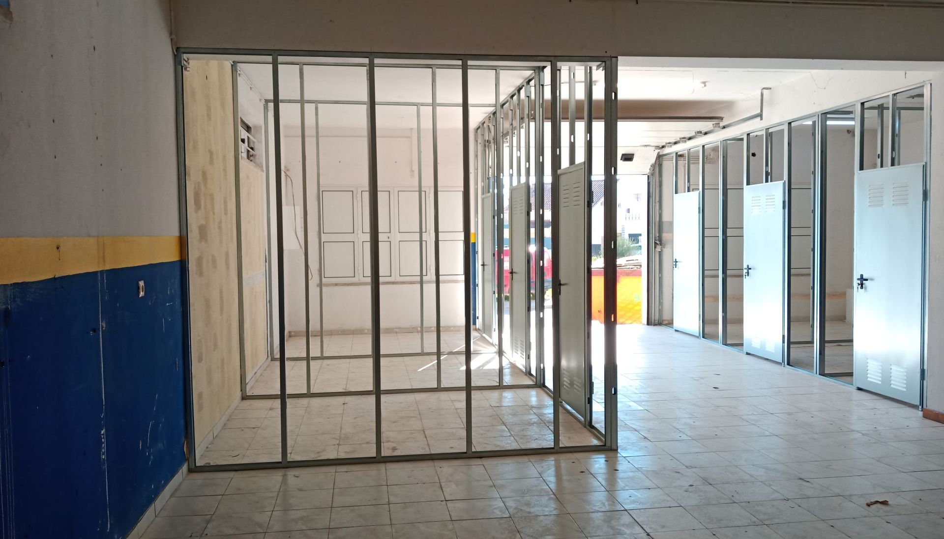 Vista interna de um edifício em construção. Estrutura metálica para paredes e portas. Pintura azul e amarela em uma parede existente.