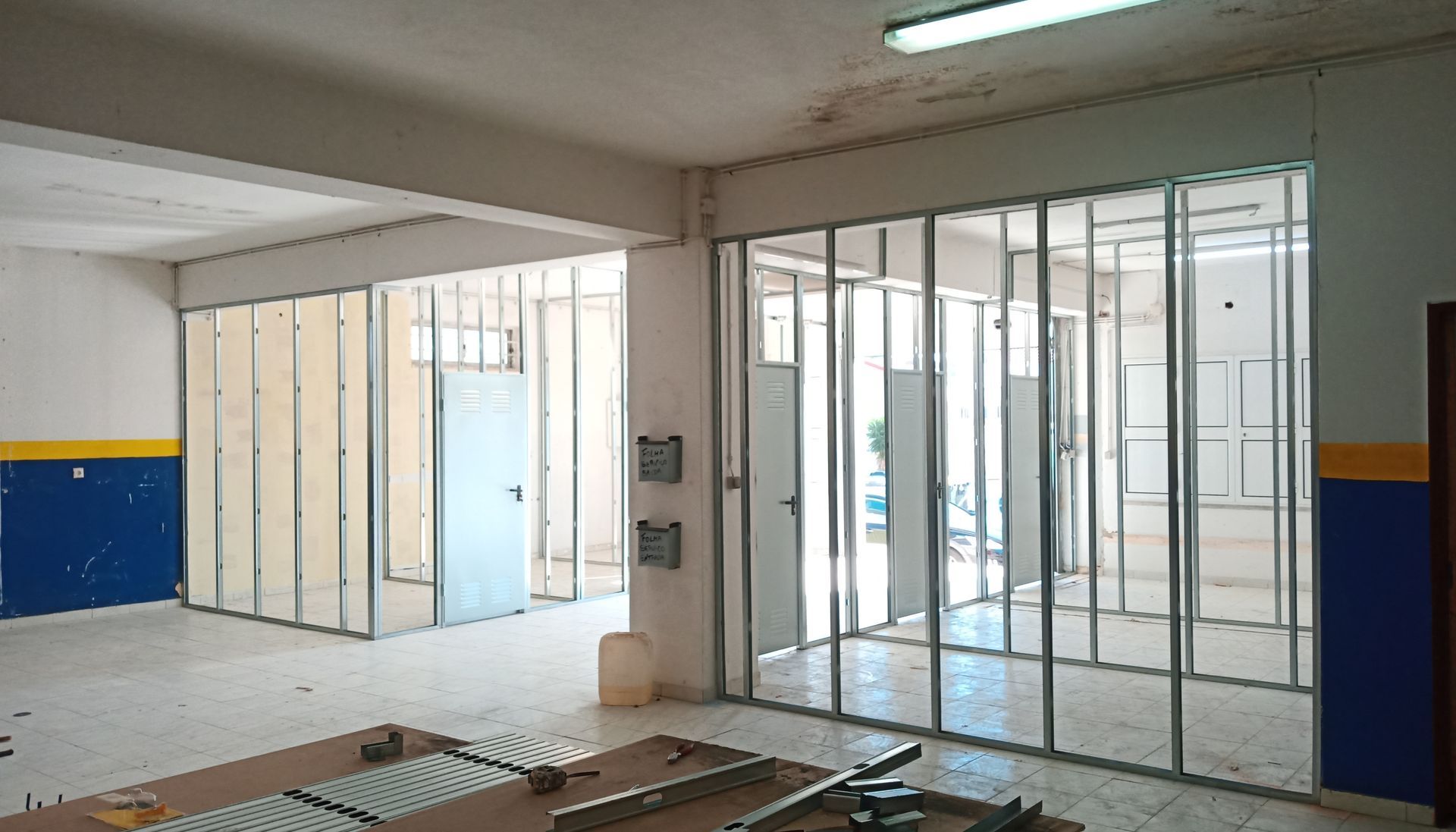 Quarto interior em construção, estrutura de paredes com montantes metálicos, portas visíveis, pintura azul/amarela nas paredes.