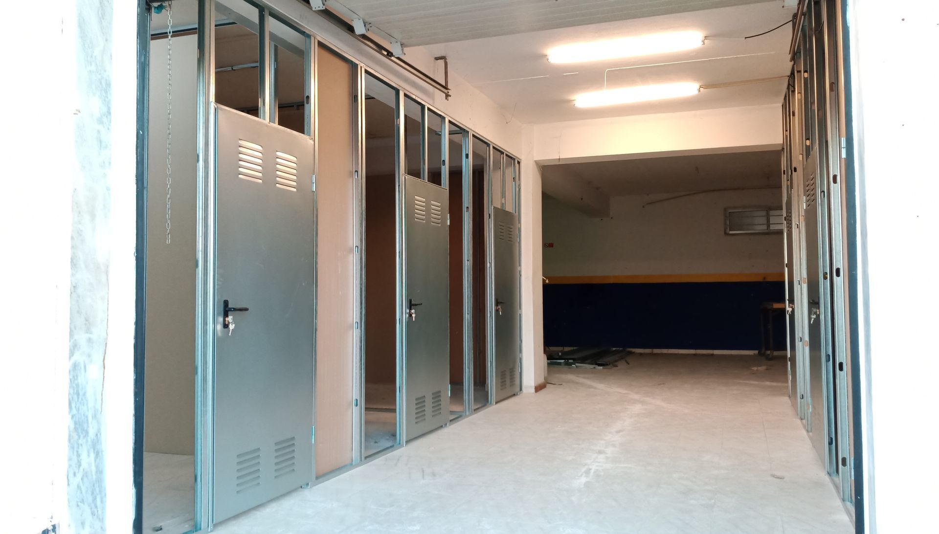Armários de metal para armazenamento em um corredor, iluminados por luzes fluorescentes. Paredes e piso de cimento.
