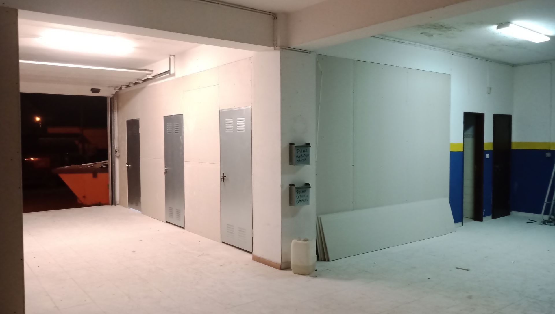 Vista interior de uma oficina com paredes brancas, três portas de metal e uma porta aberta para o exterior.