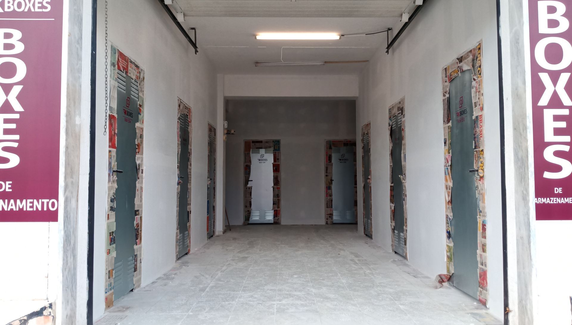 Um corredor aberto com várias portas. As portas têm molduras coloridas e estampadas. As paredes são brancas e o chão é cinza.