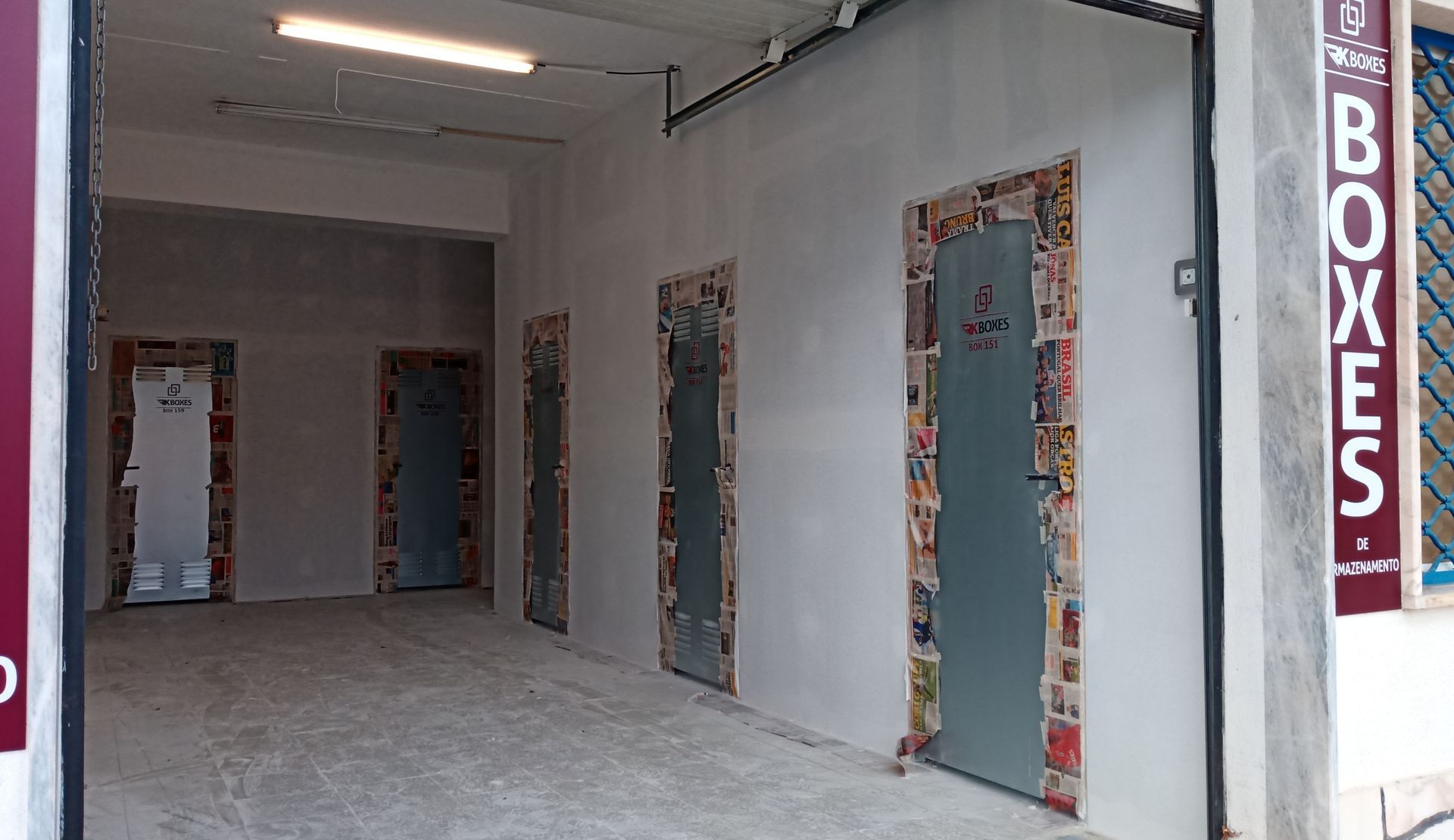 Vista interna de um depósito com várias portas revestidas com jornal nas bordas. As paredes são cinza e o piso de concreto.