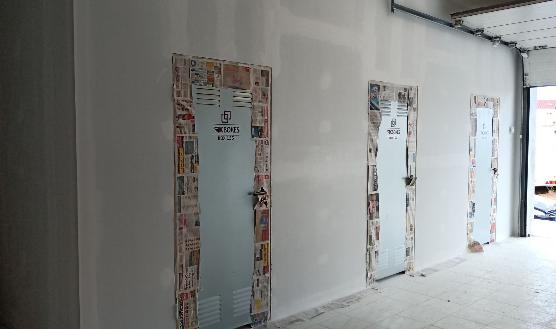 Três portas de metal, cada uma com uma borda decorativa feita de jornal, em um corredor de paredes brancas.
