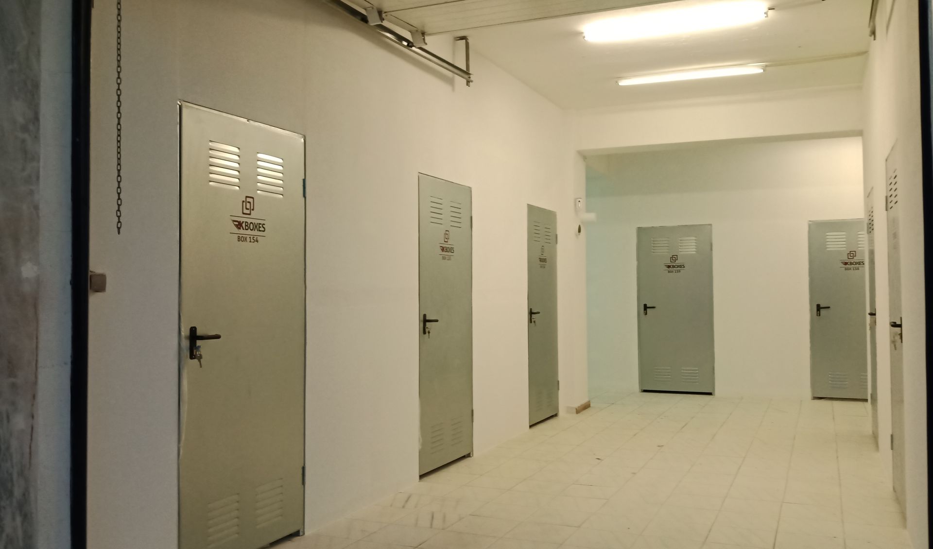 Corredor com várias portas de armários de metal cinza fechadas. Paredes e teto brancos, iluminação fluorescente.