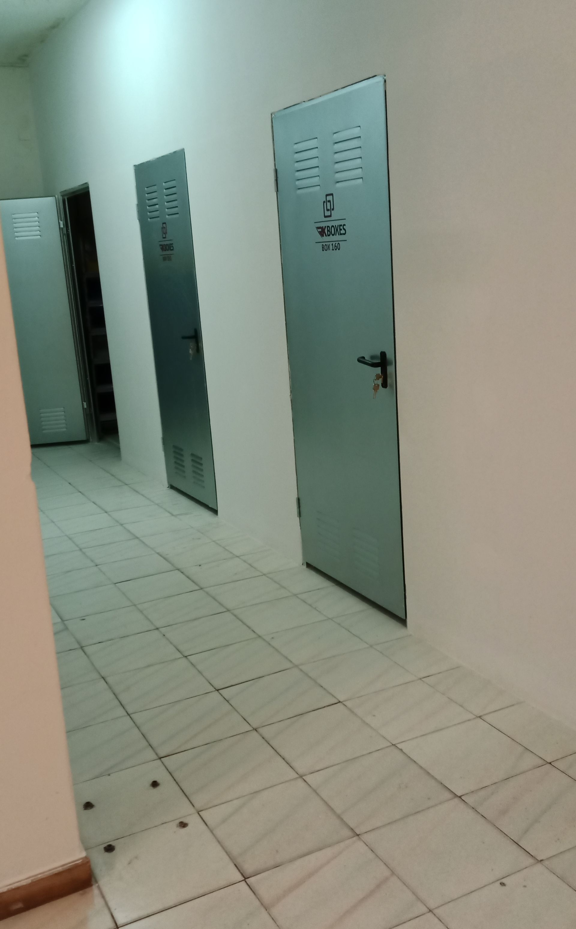 Um corredor com três armários de metal e piso de azulejo. As paredes são brancas com um tom verde claro.