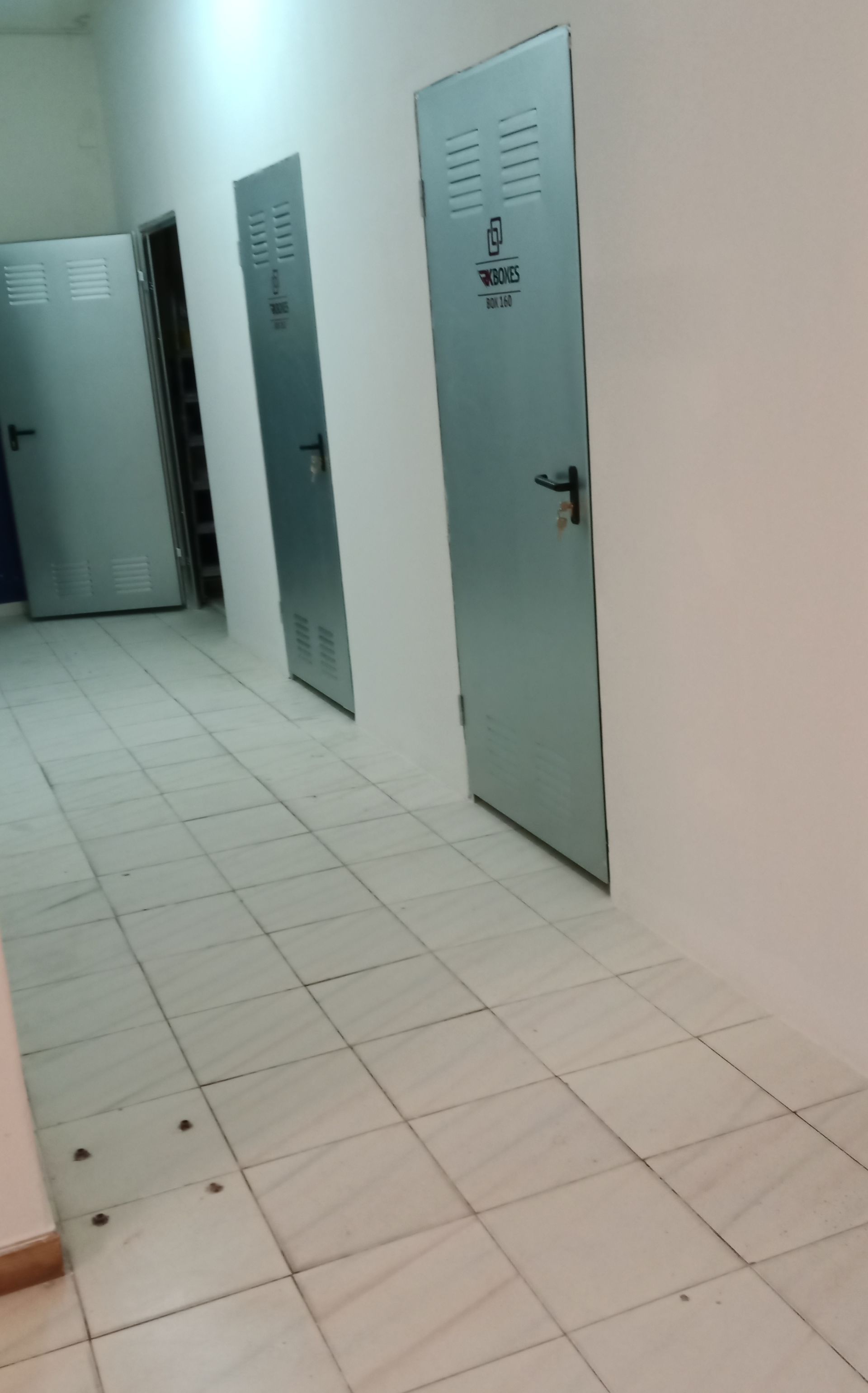 Corredor com paredes brancas, portas de metal cinza numeradas de 1 a 3 e piso de azulejo.