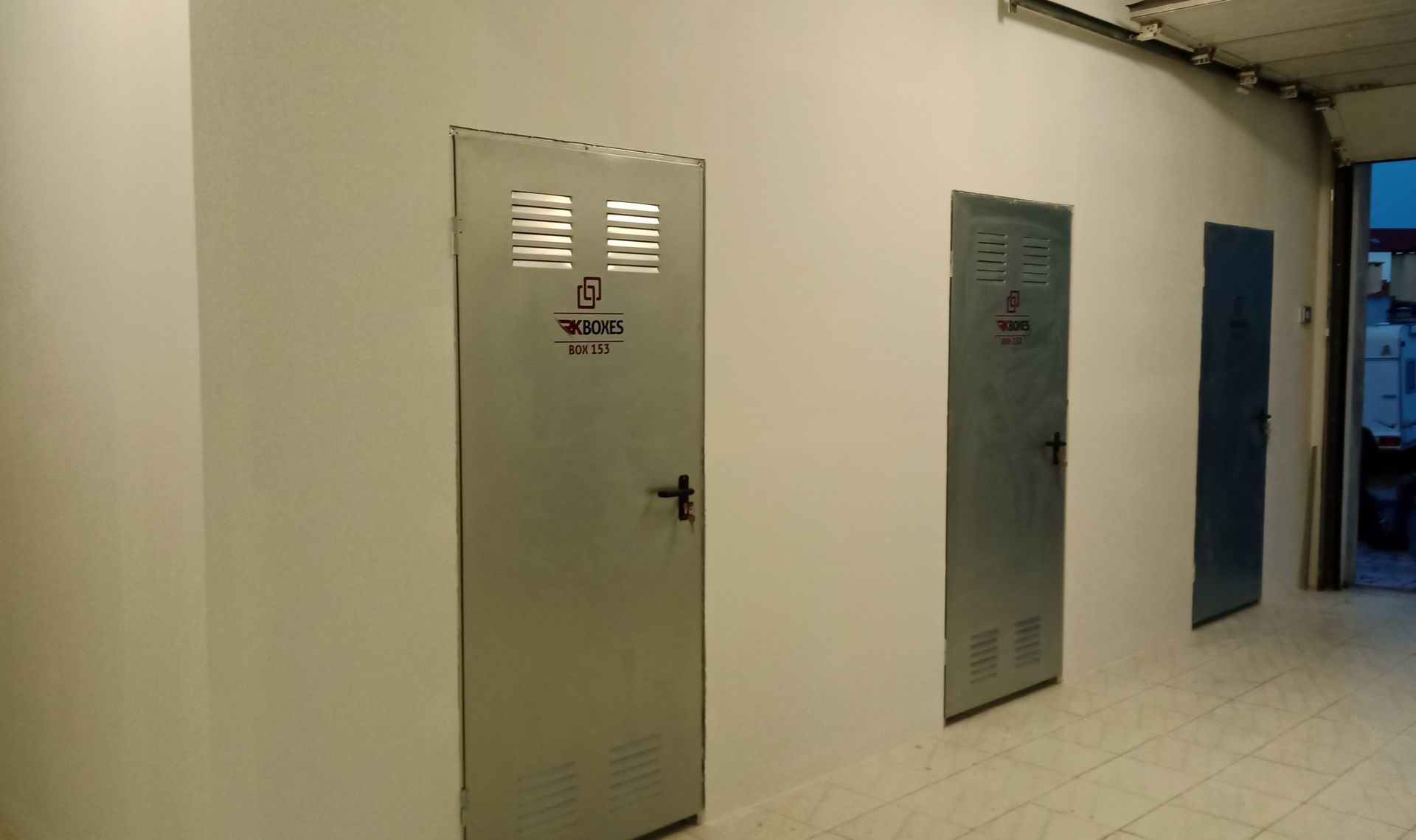 Três portas de metal em uma parede branca em um corredor. A primeira porta possui aberturas de ventilação e uma maçaneta.