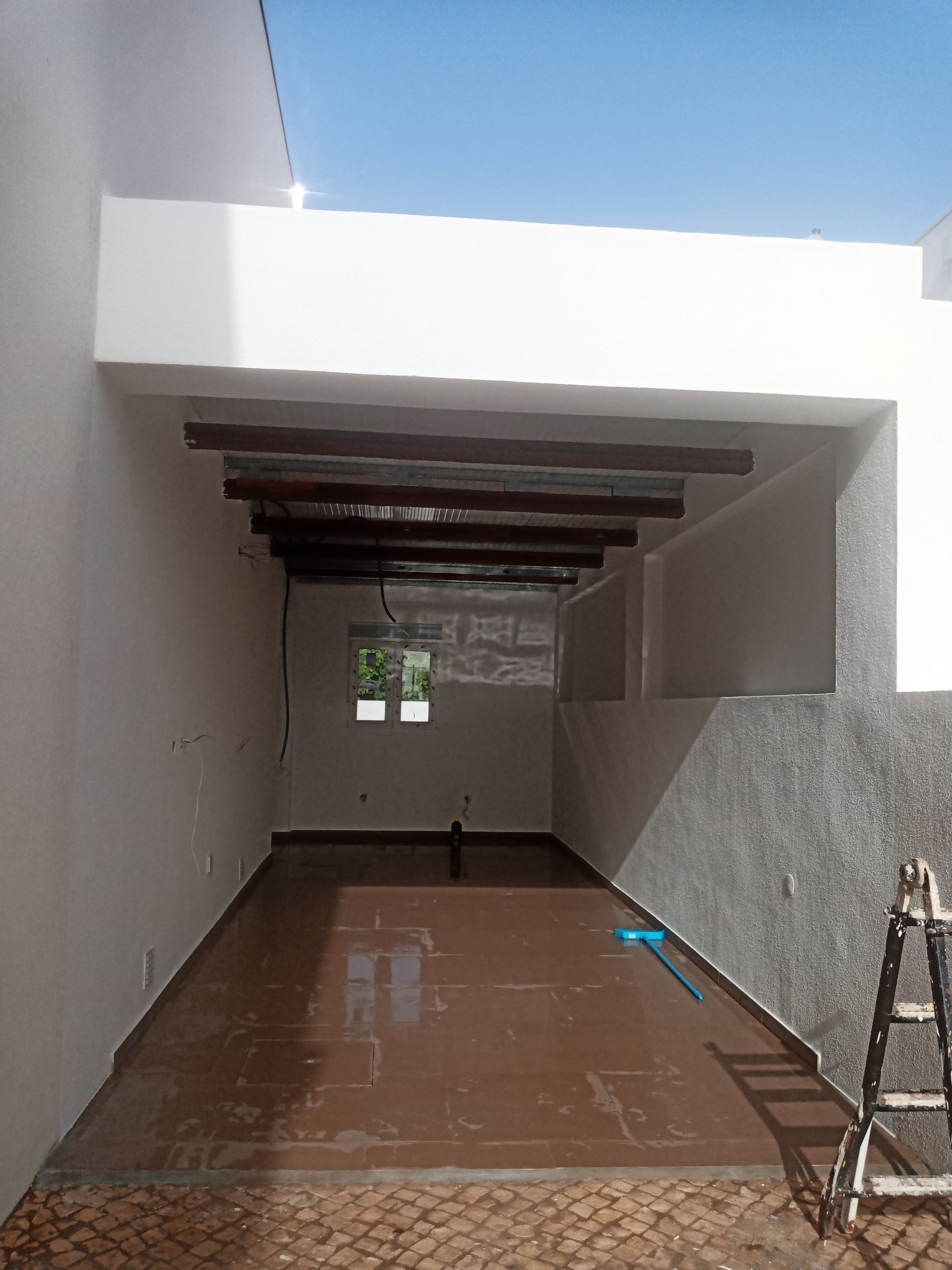 Um espaço externo coberto com piso de azulejo marrom, paredes brancas, vigas de madeira escura e um vislumbre do céu.