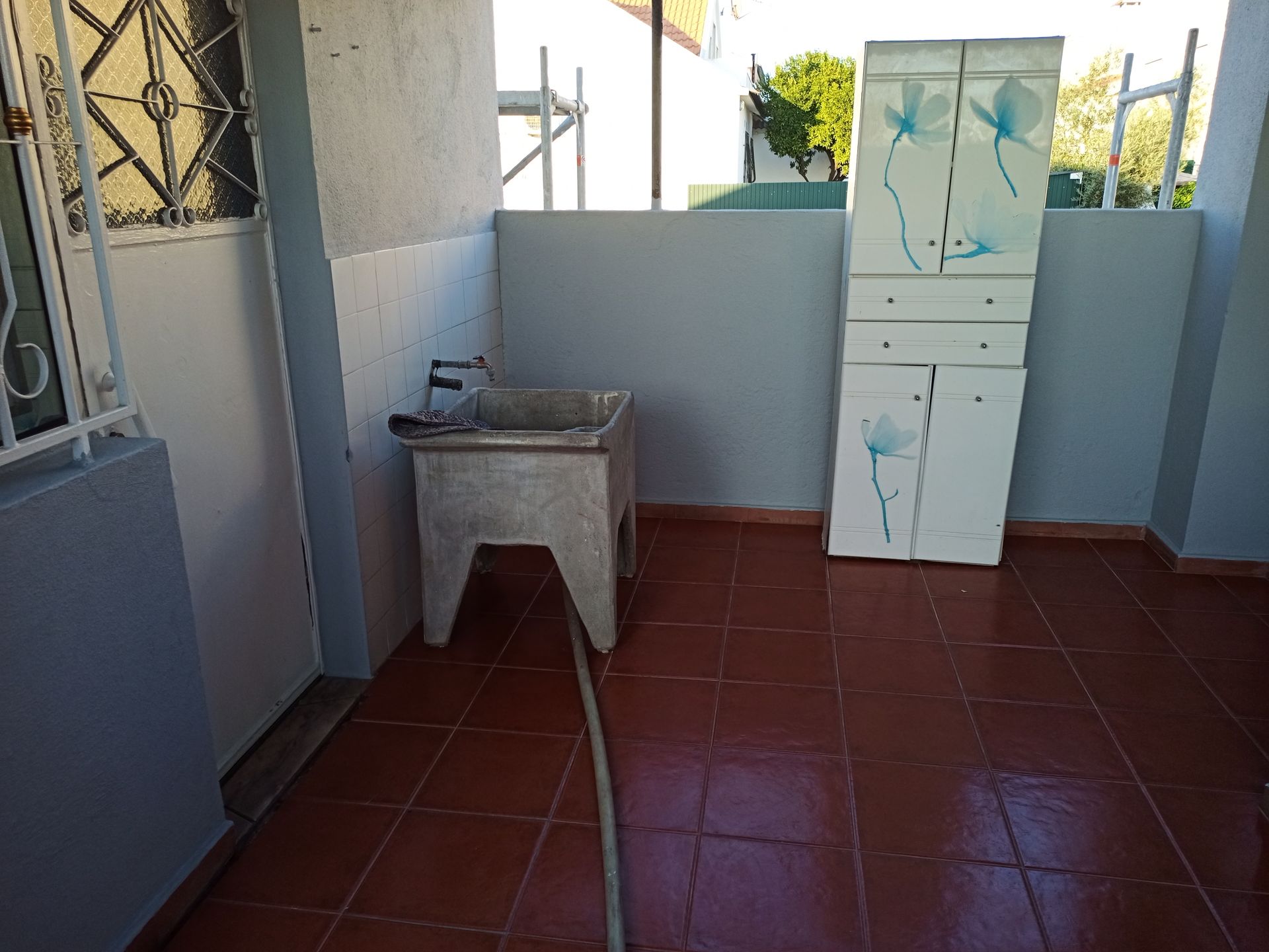 Lavanderia com pia de concreto, armários e mangueira sobre piso de ladrilho vermelho.