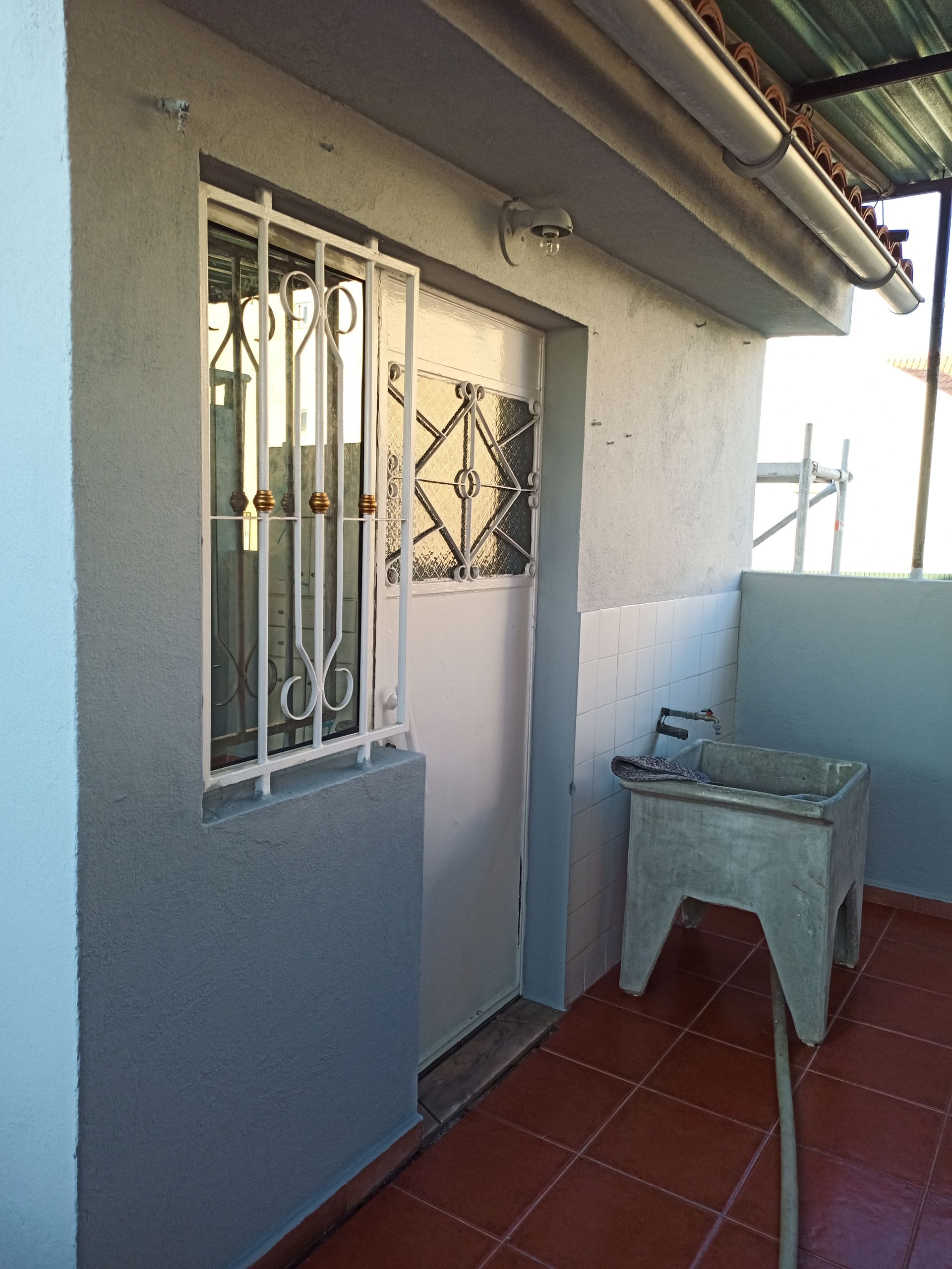 Exterior com janela, porta e lavatório. Paredes cinzentas, grades de segurança brancas, piso de ladrilho vermelho.