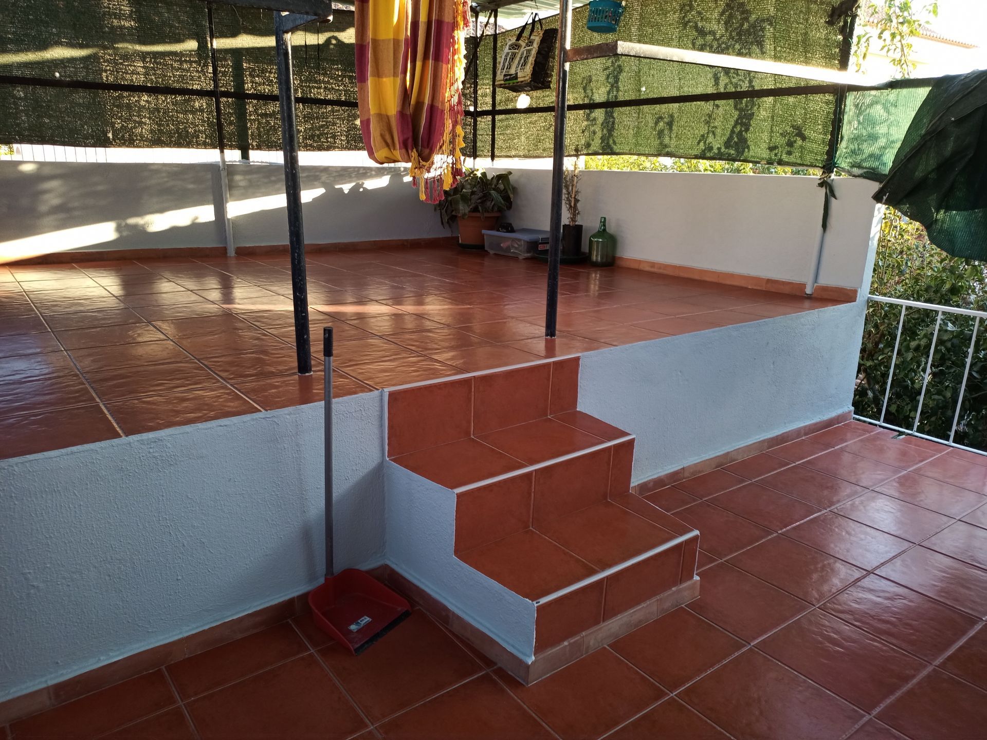 Terraço com piso de ladrilho vermelho, paredes brancas e três degraus. Há uma vassoura e uma rede.