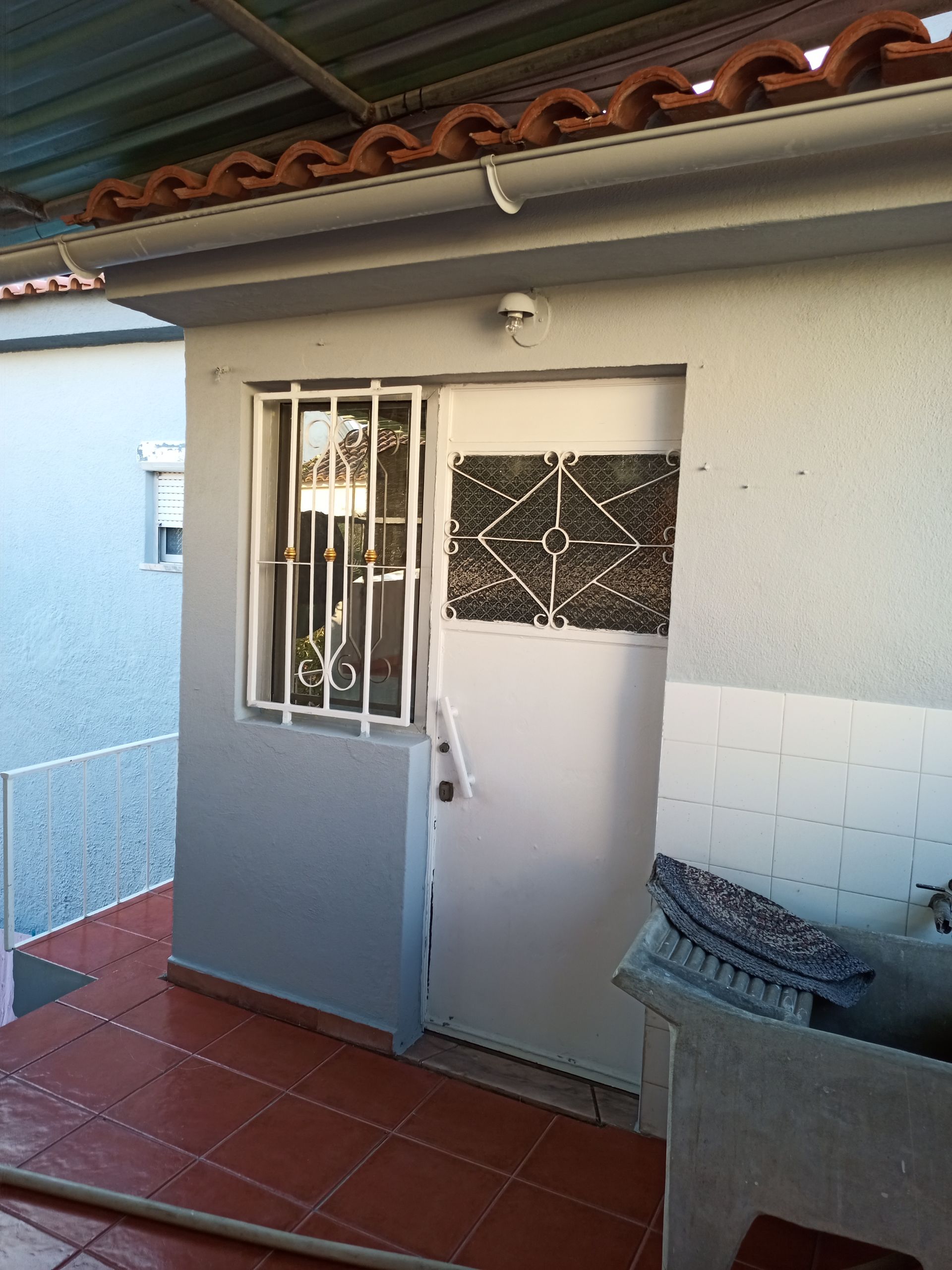 Exterior de um edifício com porta branca e janela gradeada. Piso de ladrilho vermelho e telhado de metal.