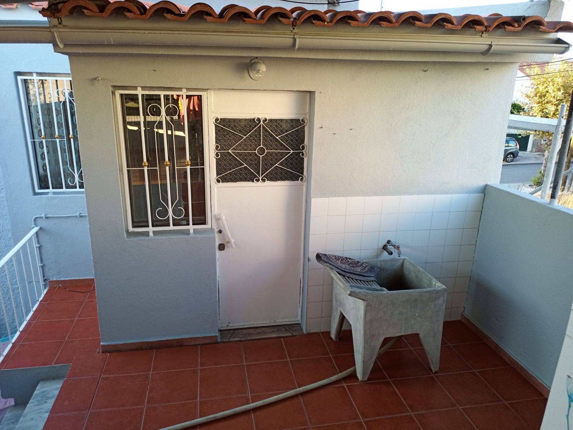 Exterior de uma pequena estrutura branca com piso de ladrilho vermelho e pia de concreto. Porta e janela com grades.