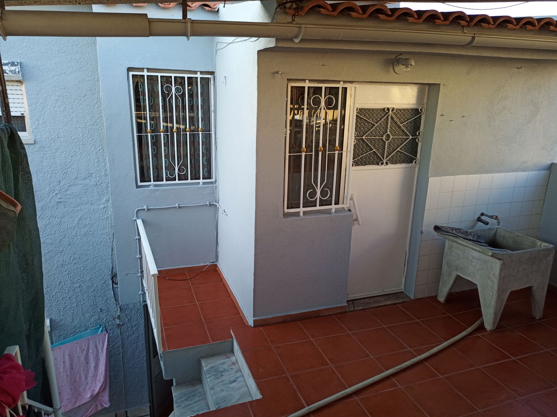 Vista exterior de um pequeno pátio com parede pintada de azul, porta, janela gradeada e pia de serviço.