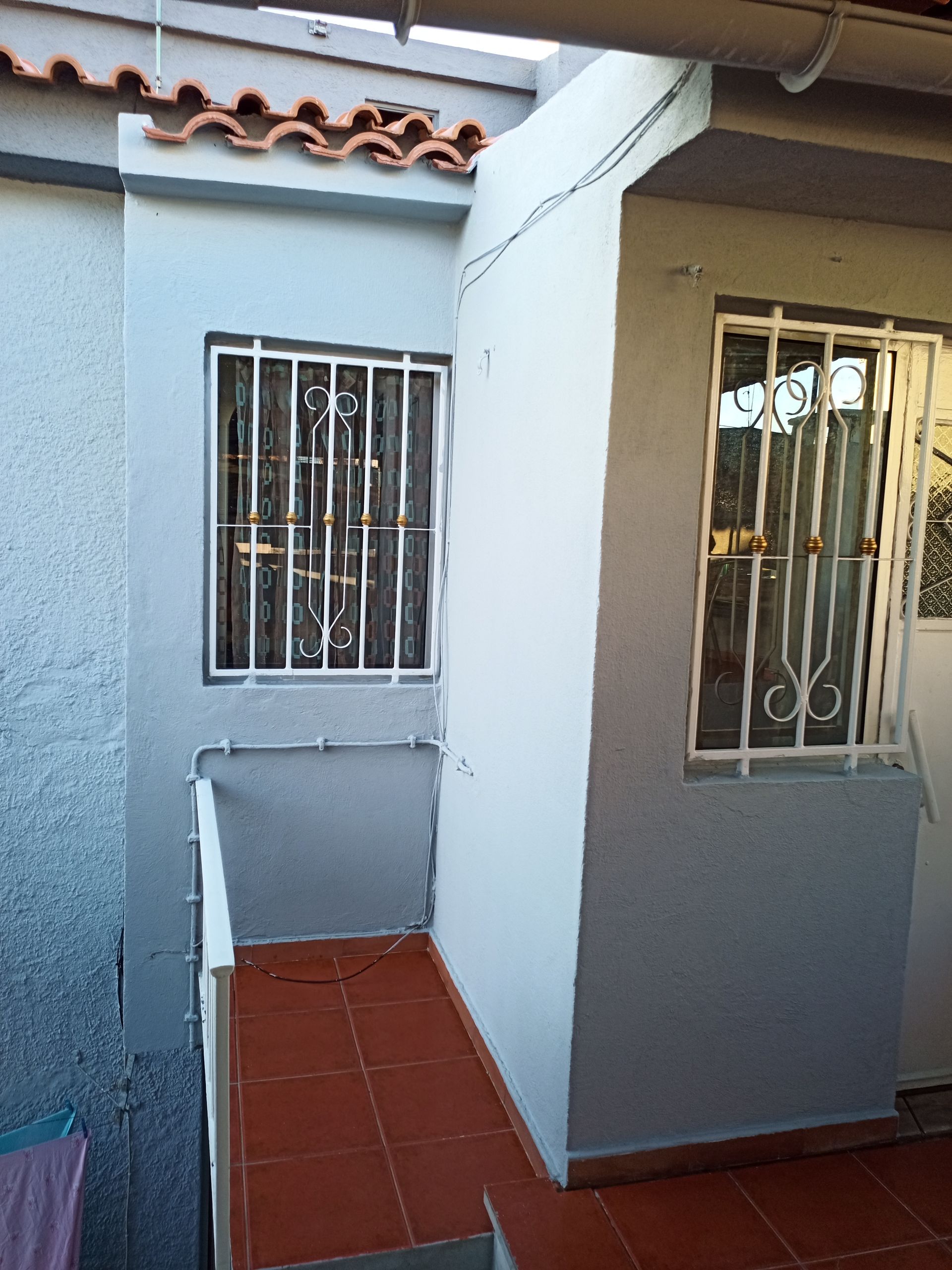 Vista exterior de um pequeno edifício com janelas gradeadas, pintado de cinza, com piso de ladrilho vermelho.