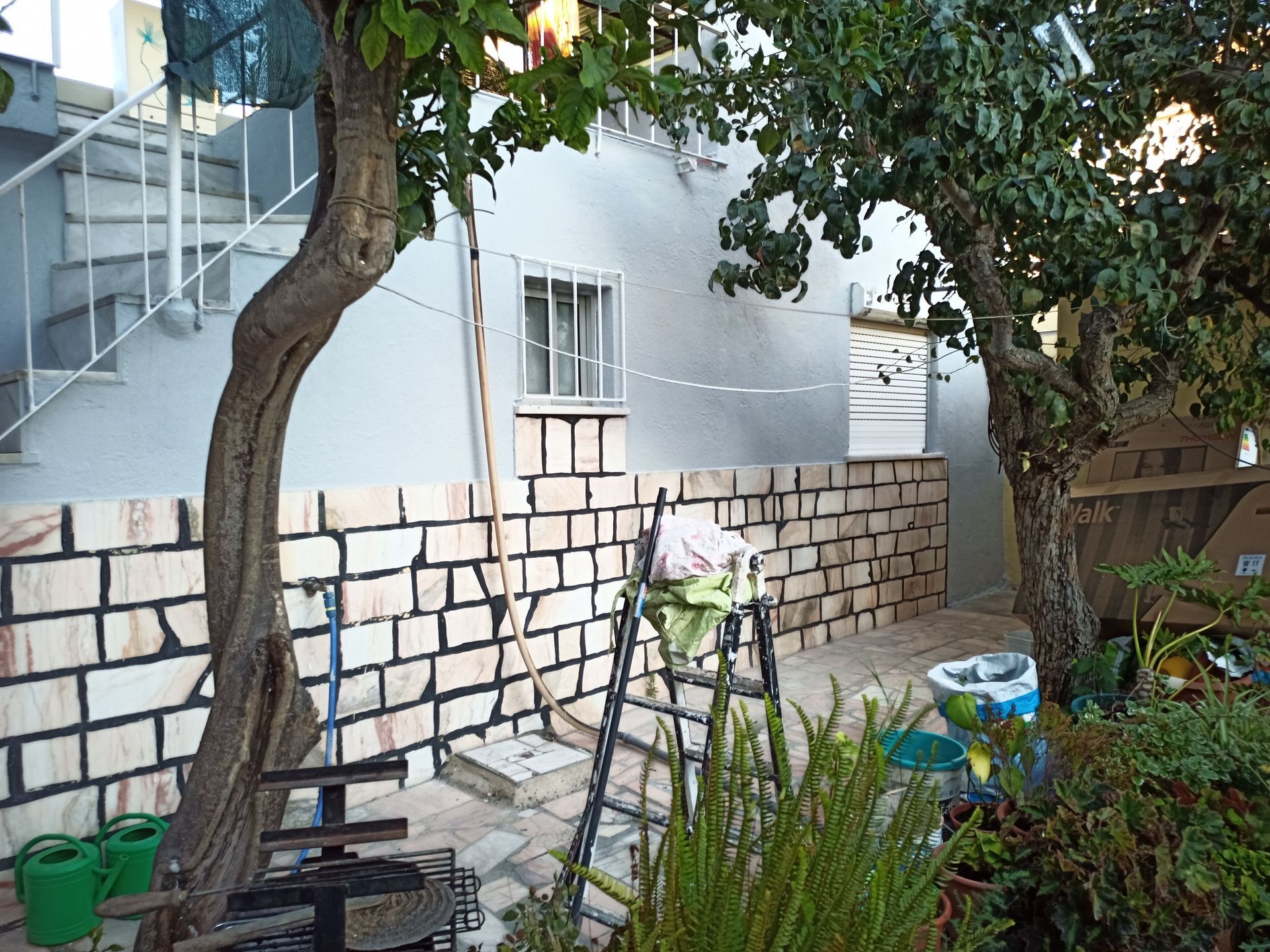 Exterior de um edifício com parede de tijolos, árvores e uma escada em espiral. Uma escada está encostada na parede.